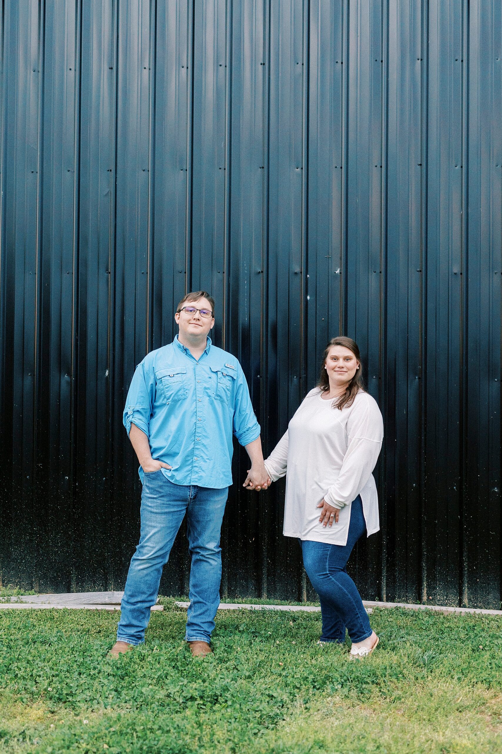 woman in ivory sweater holds hands with man in blue shirt in front of metal barn at the Farmstead
