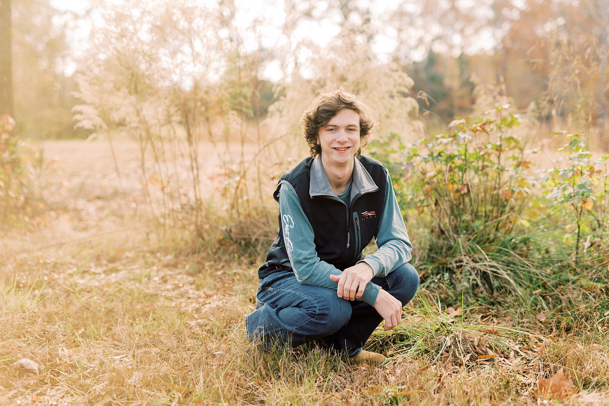 senior boy kneels in tall grass during senior portraits in Midland NC