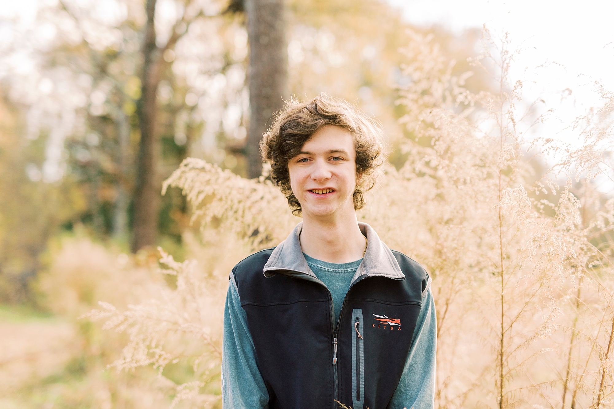 boy in black vest and blue shirt stands in tall grass during fall Midland NC senior portraits 
