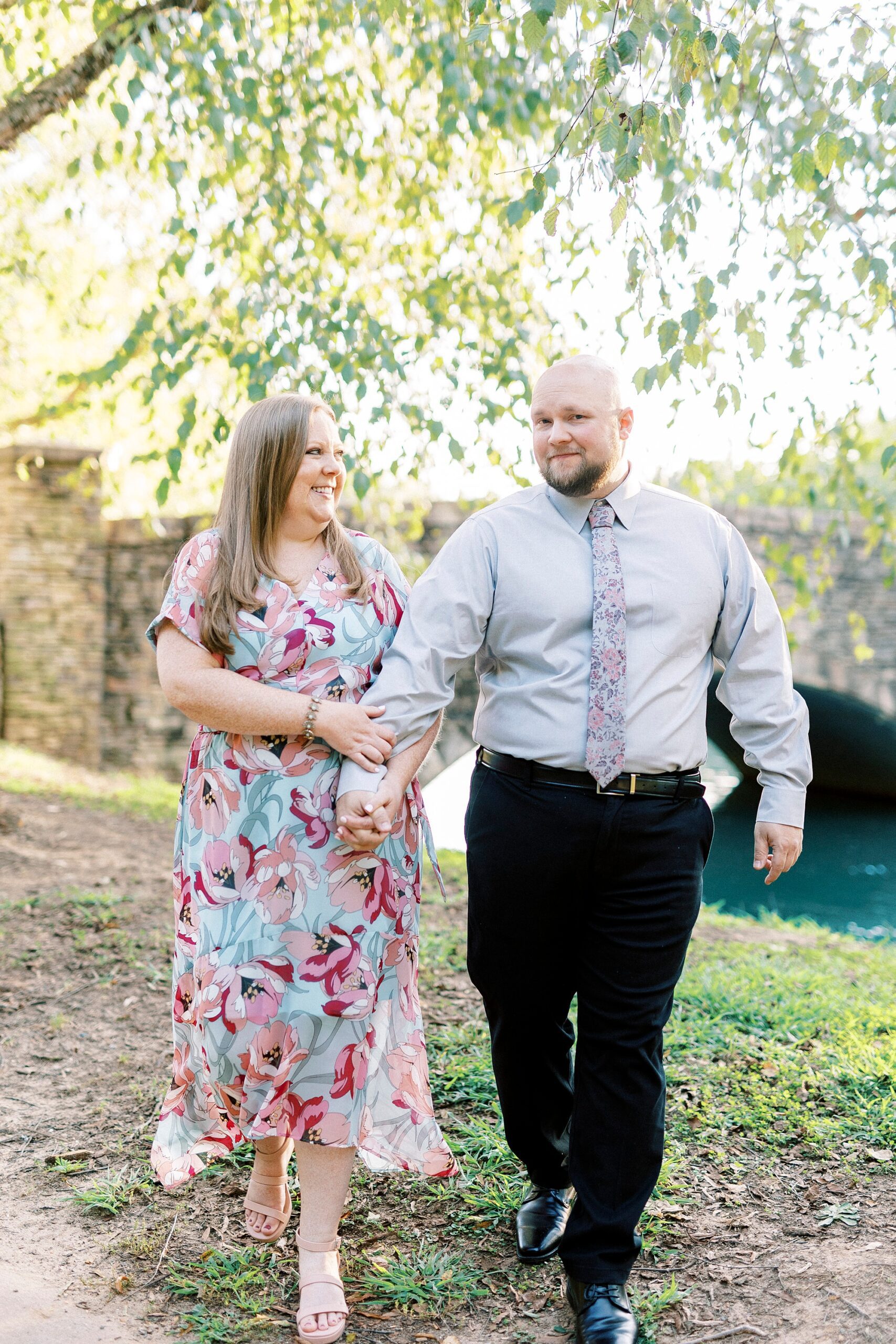 couple holds hands walking near stone bridge under weeping willow in Freedom Park