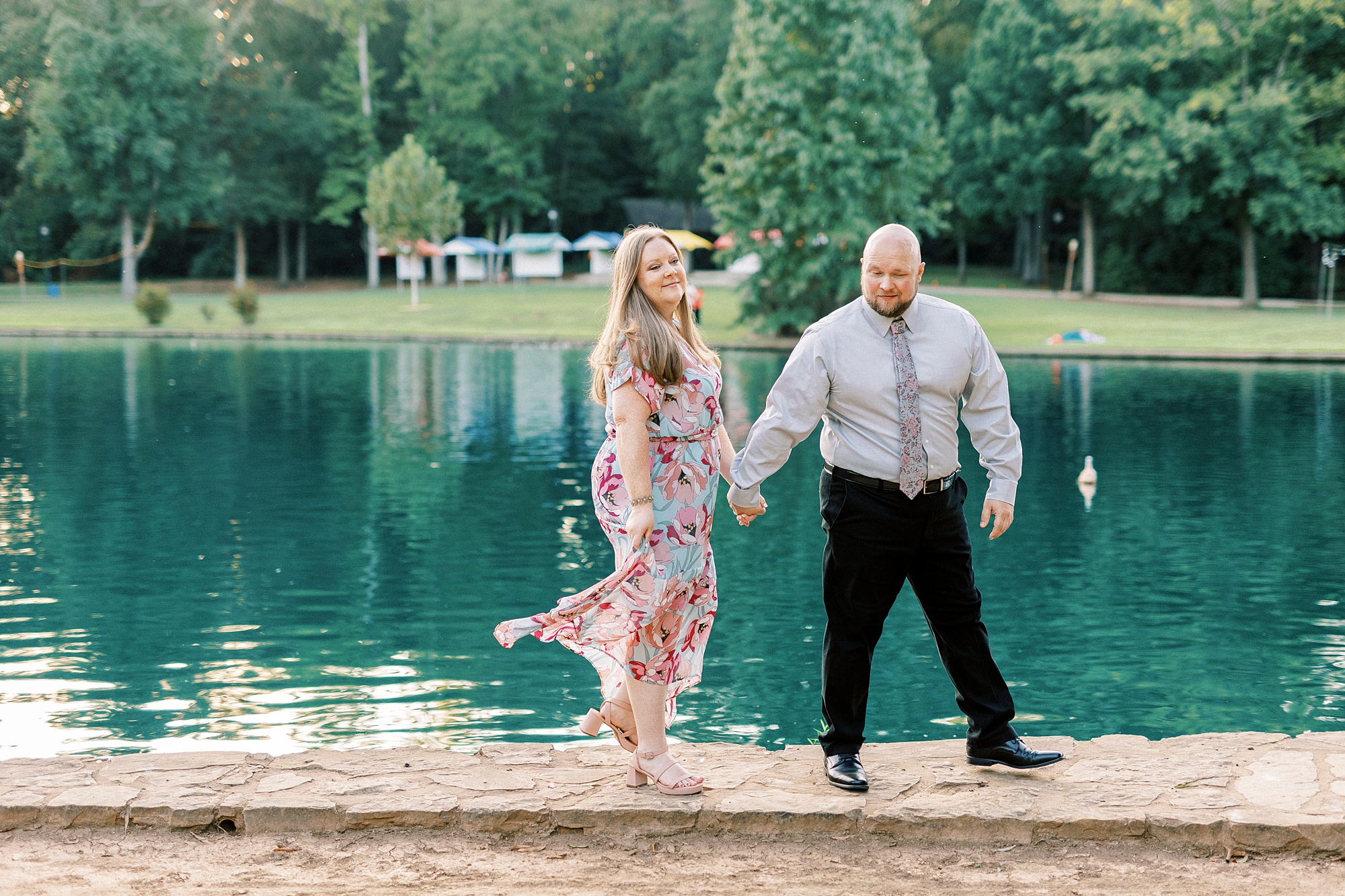 engaged couple holds hands walking on stone pathway near pond