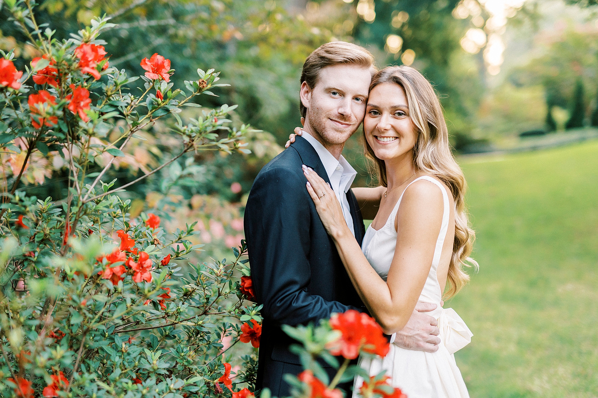 woman in white dress with bow on back hugs near red flower bush in South Carolina