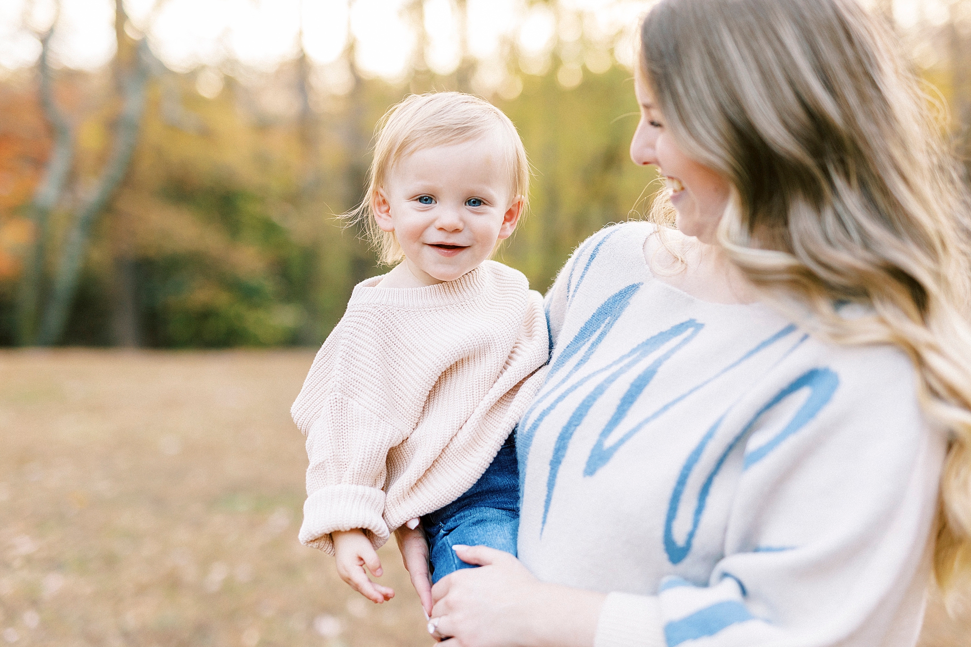 woman in cream and blue sweater dress holds blonde son on hip during photos at Baker's Creek Park