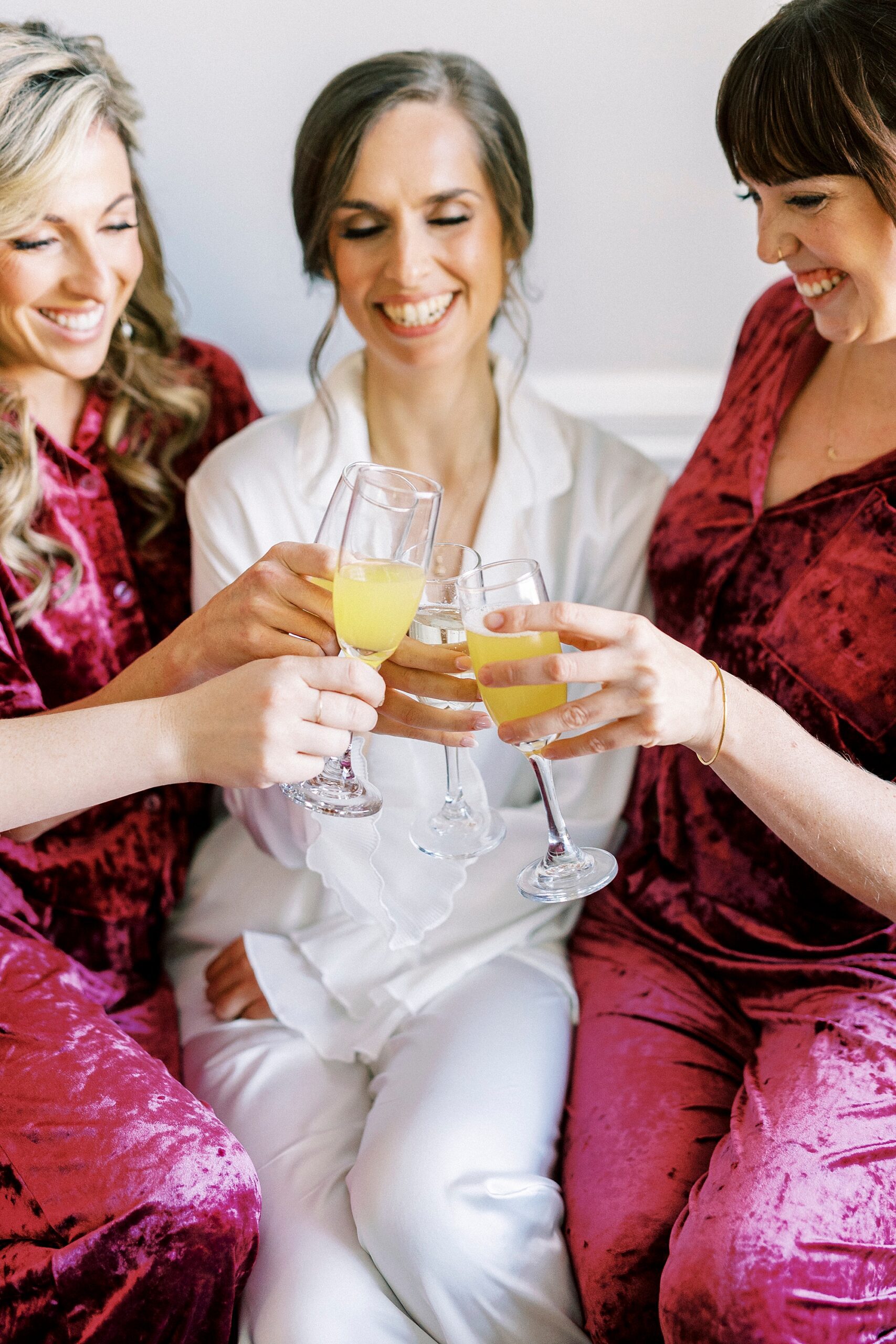 bride and bridesmaids in matching burgundy pajamas toast mimosas
