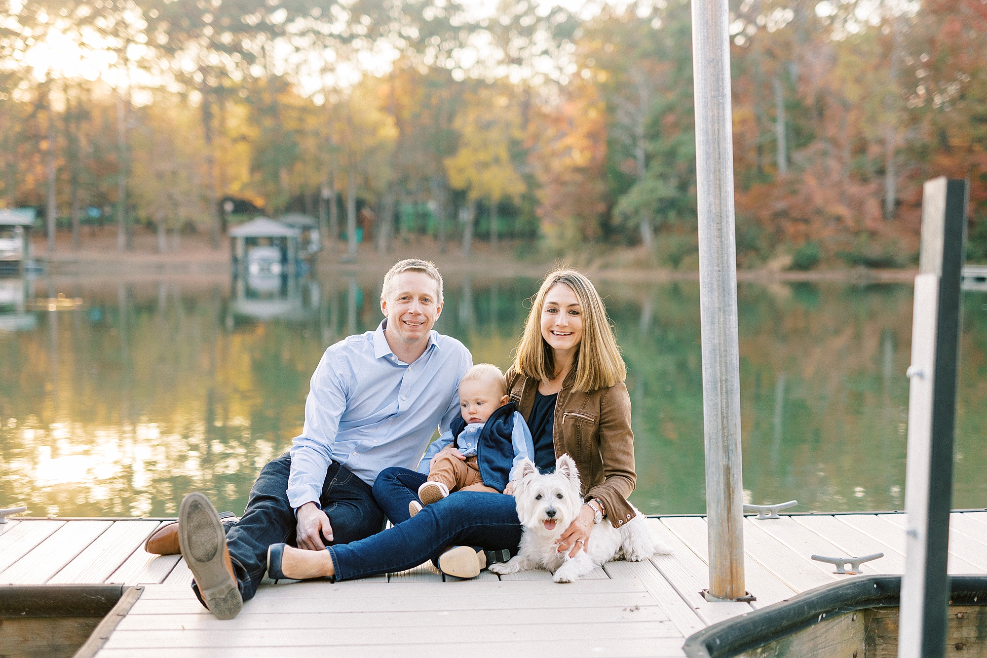 parents sit on dock holding son and white dog between them in front of lake