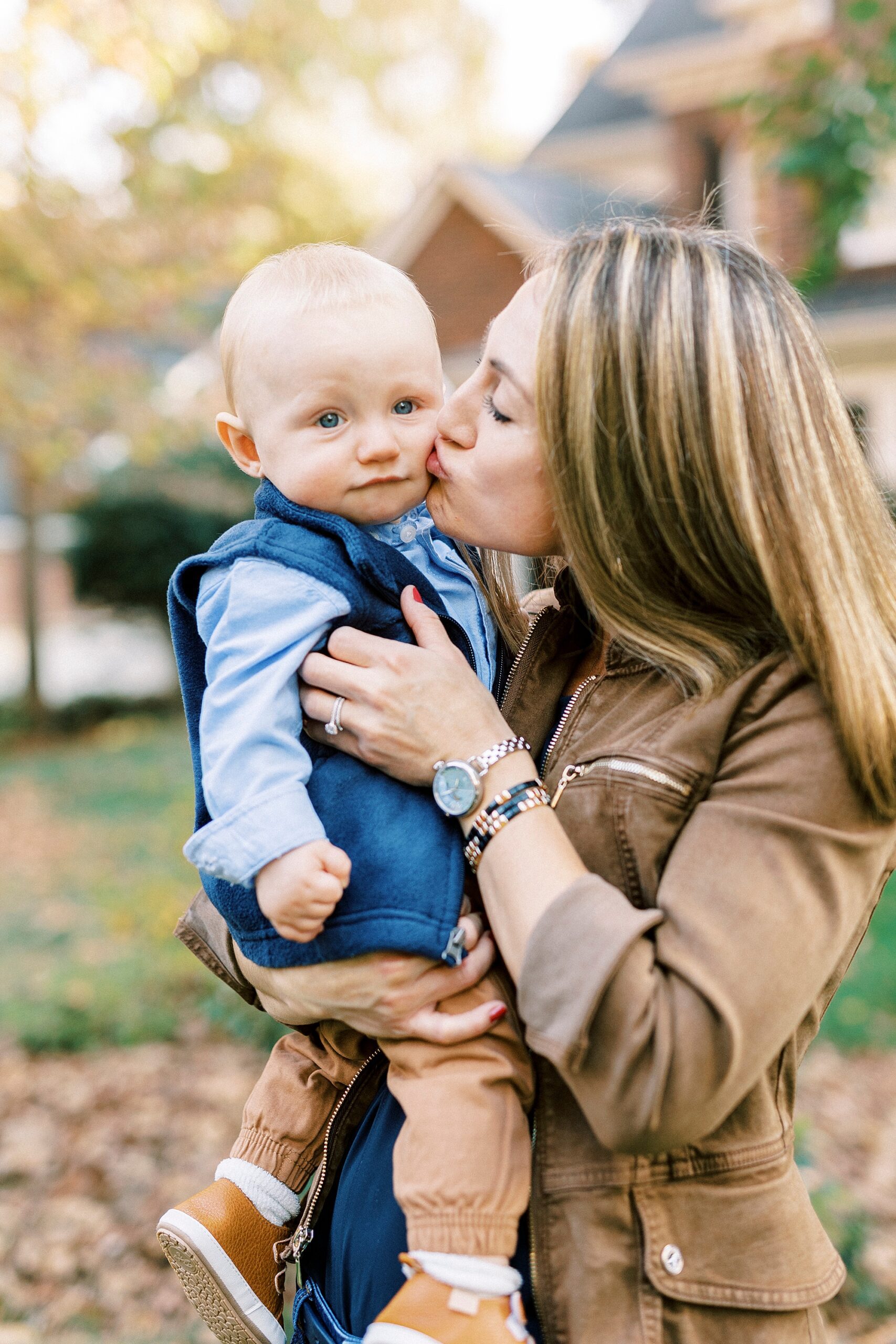 mom in brown jacket leans to kiss son's cheeks in front of home in Charlotte NC