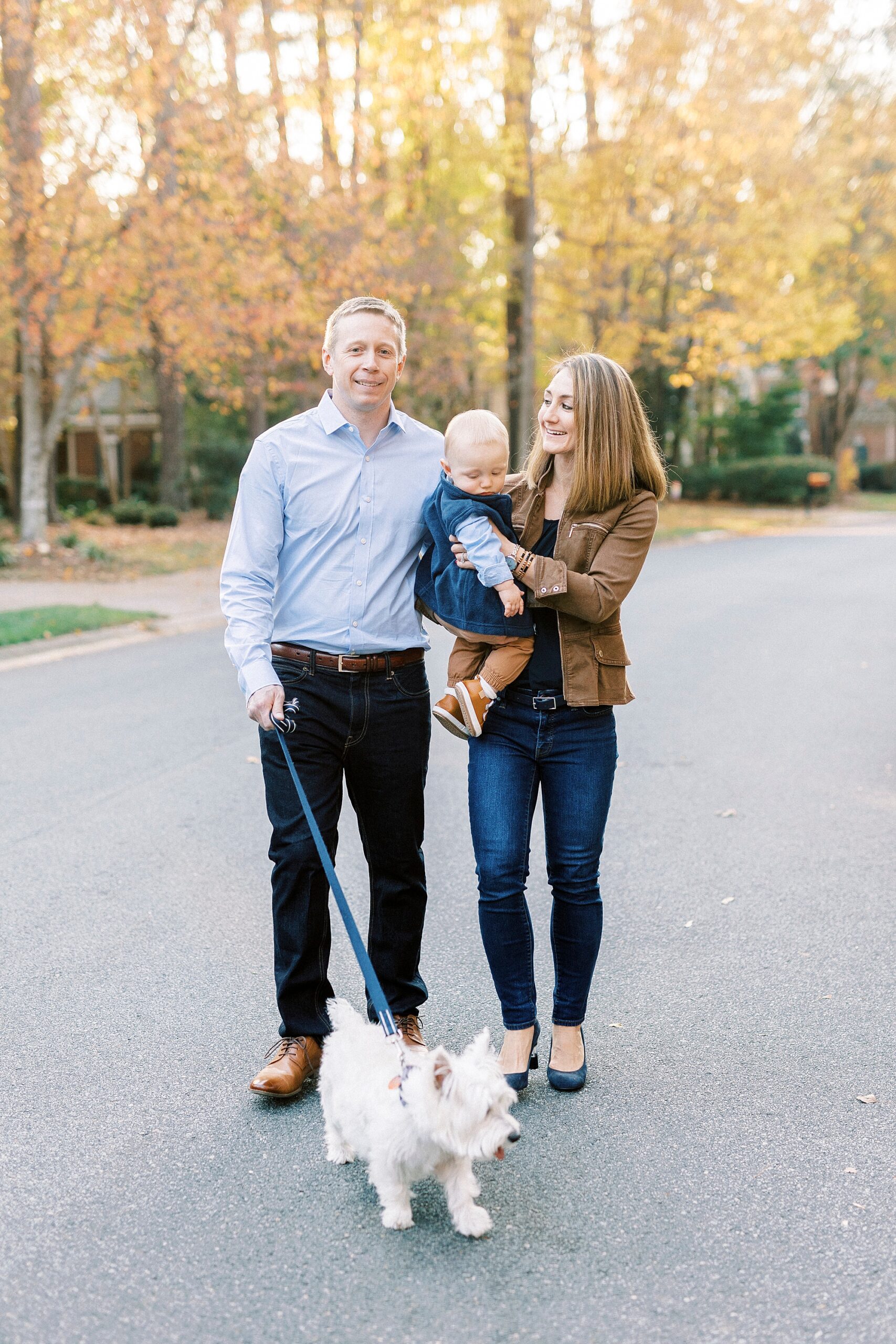 mom walks with husband while holding son on hip and he walks dog outside home