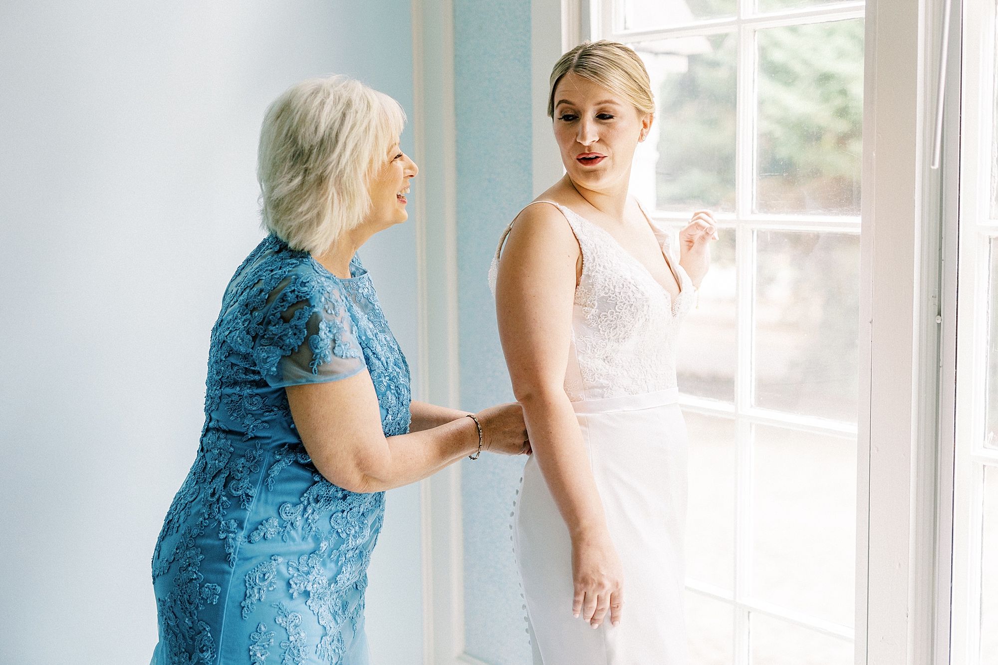 mother in blue dress buttons up wedding gown for bride looking out window