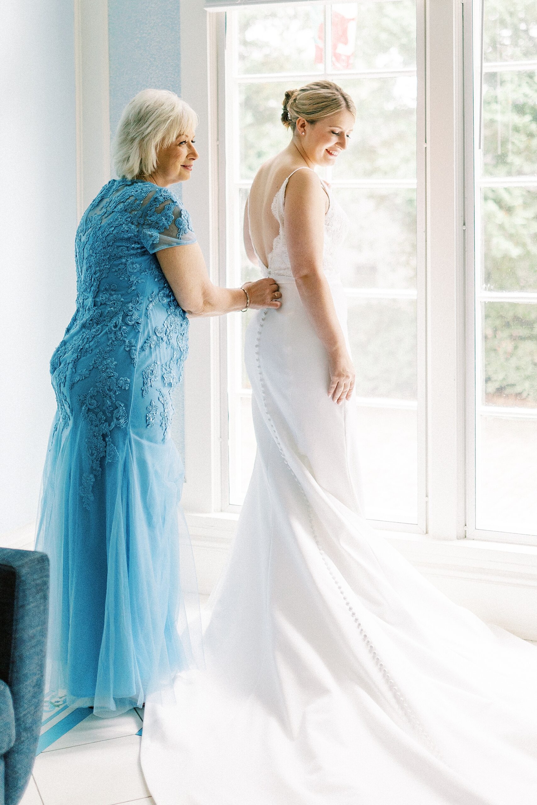 mother buttons up wedding gown for bride looking out window