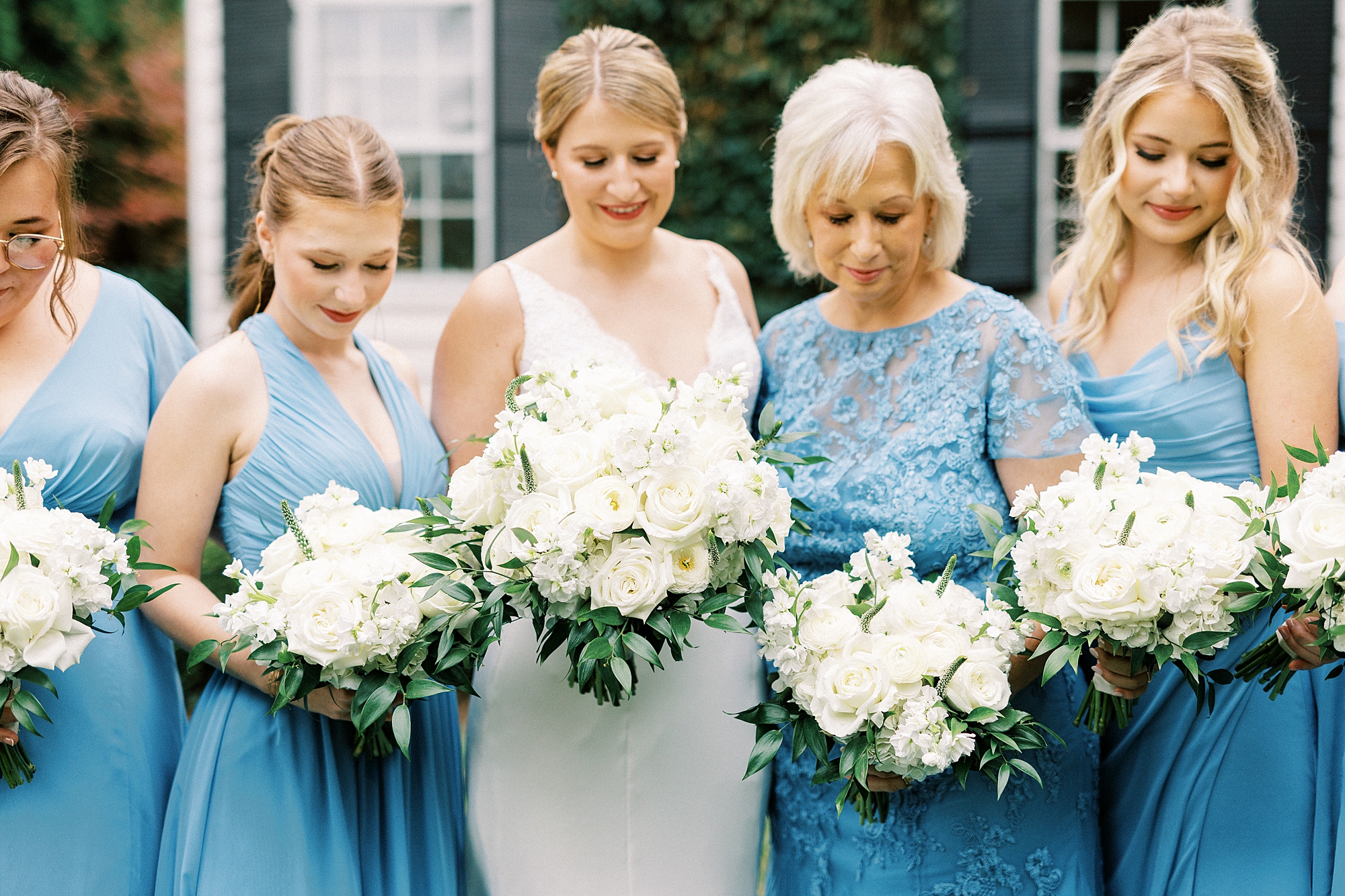 bride looks down at bouquet of ivory flowers surrounded by bridesmaids in light blue gowns