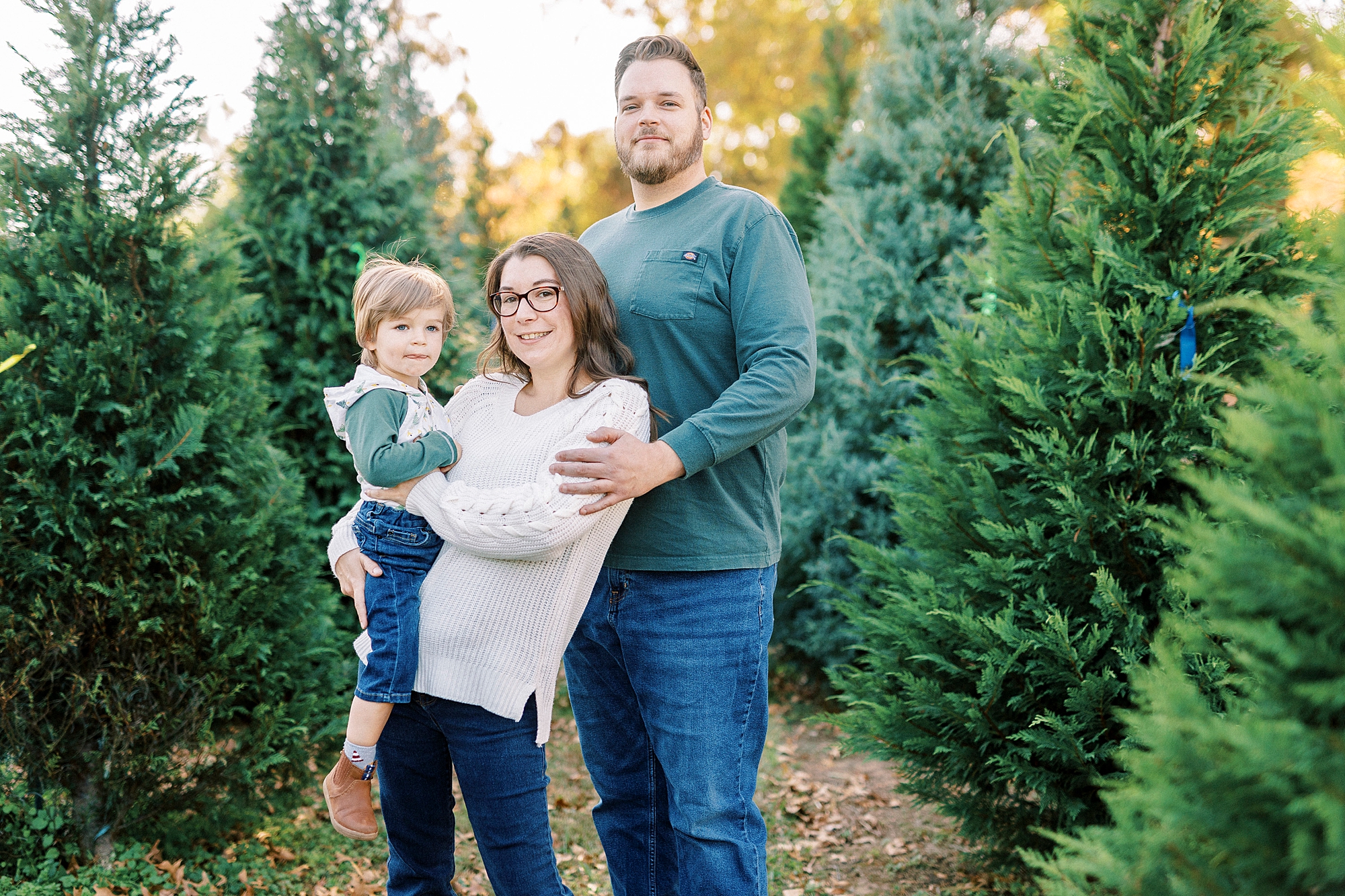 parents hug toddler between trees at Almond Christmas Tree Farm mini sessions