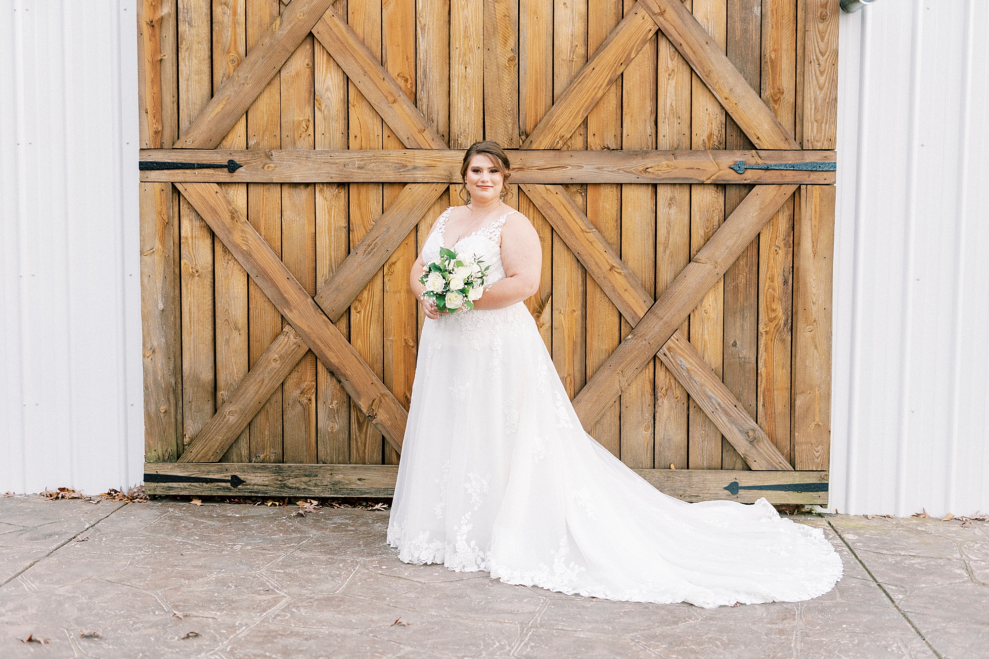 bride stands holding bouquet of white roses and baby's breath in front of wooden doors