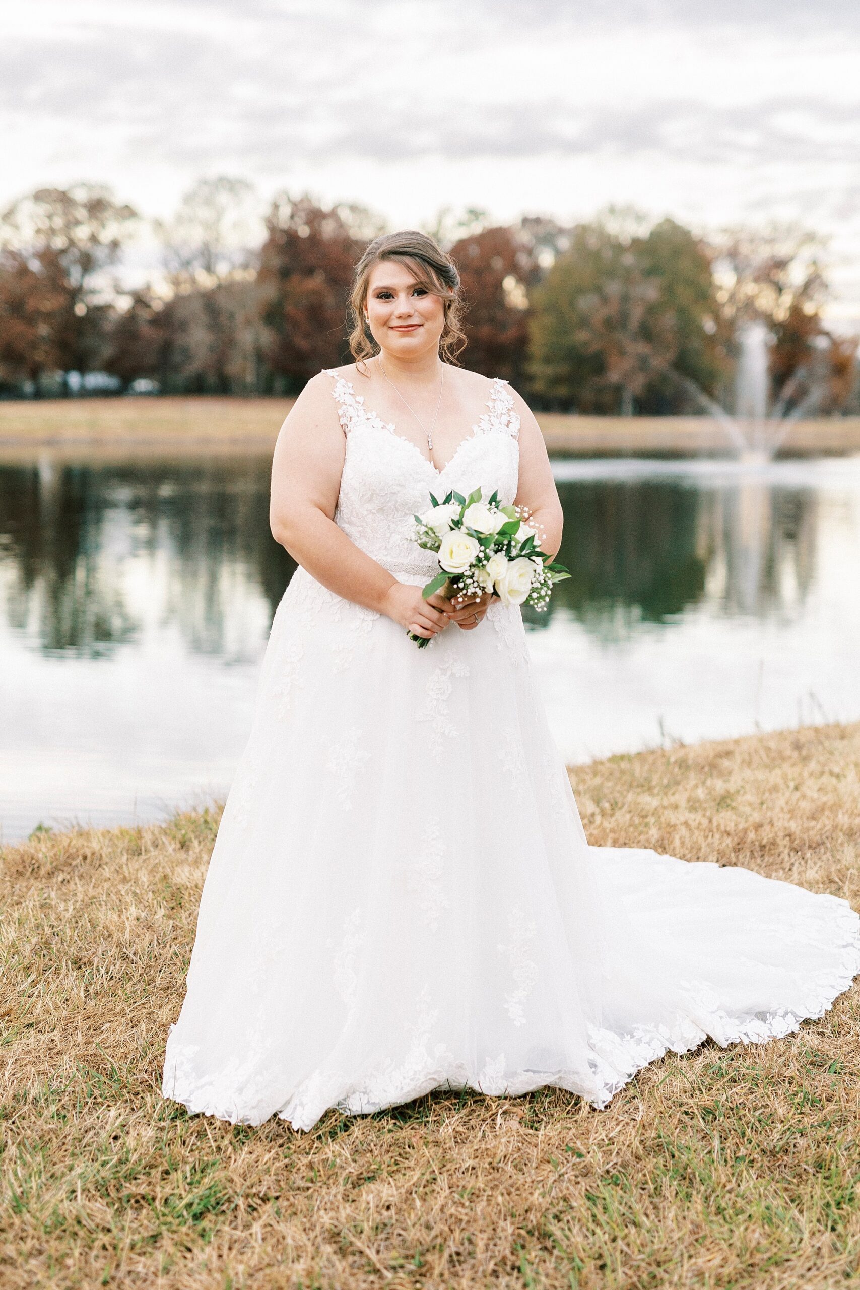 bride stands holding bouquet of white roses and baby's breath by pond at the Farm at Brusharbor