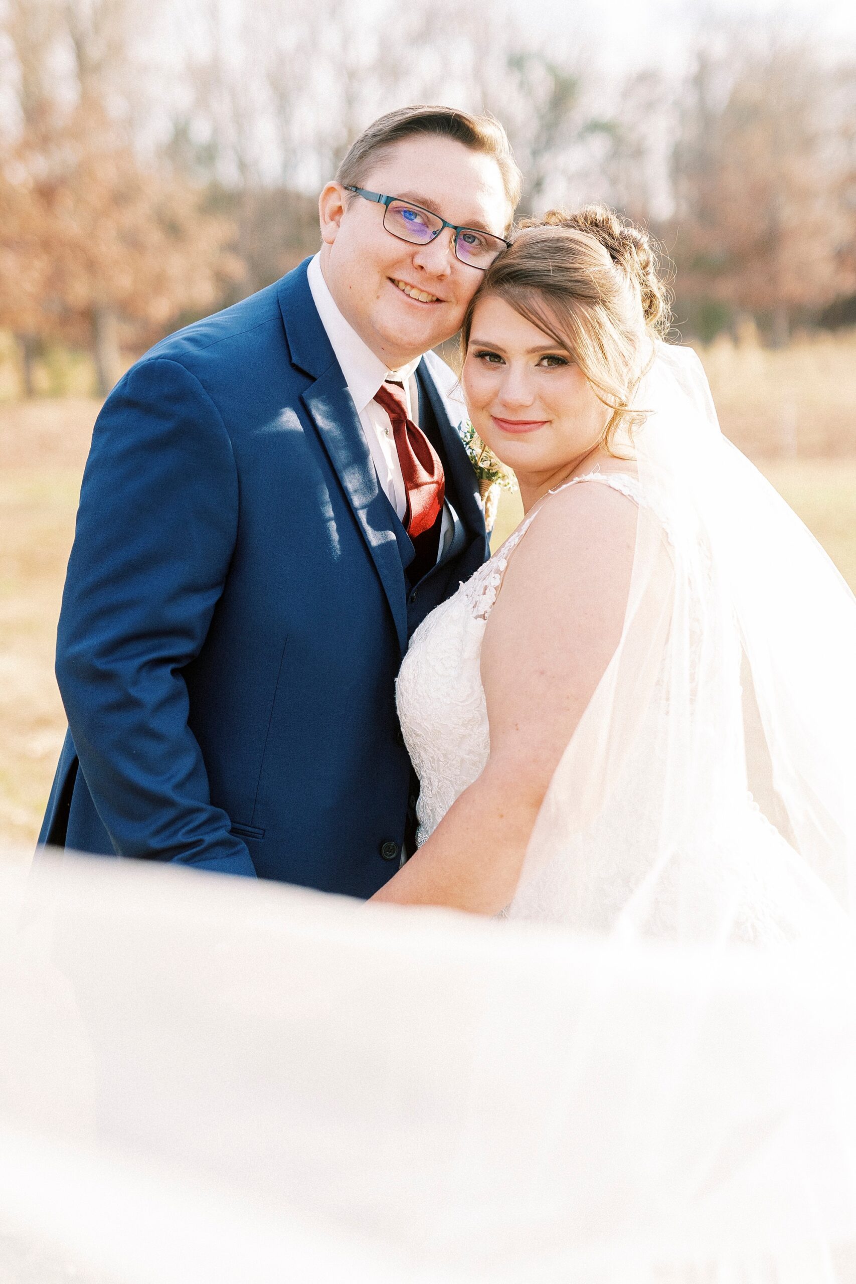 bride and groom lean heads together during fall wedding portraits with veil around them 