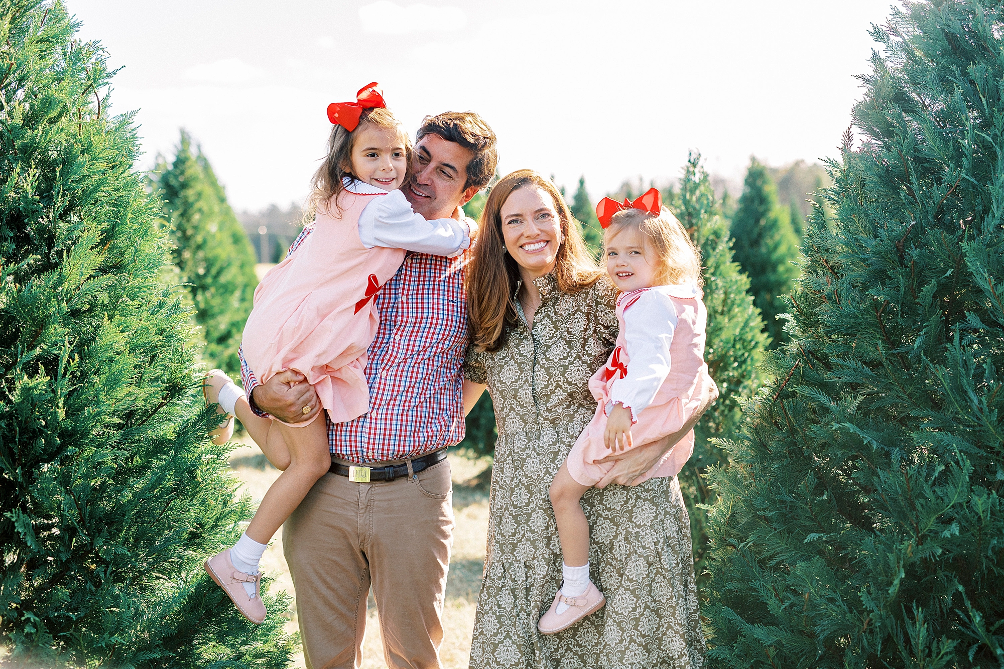 couple holds two daughters in pink and red dresses between Christmas trees