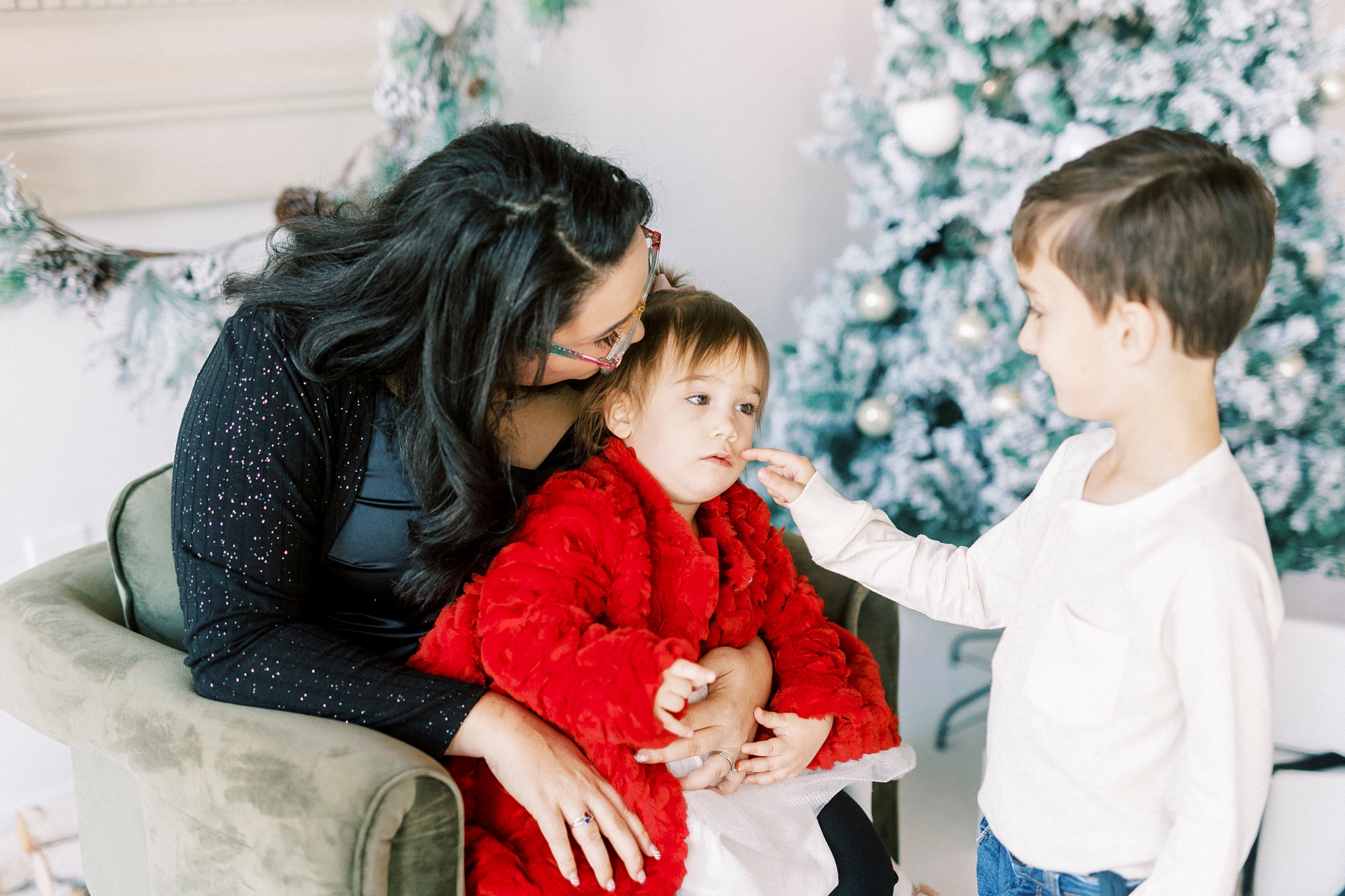 son leans to touch sisters face while she sits with mom during portraits in Downtown Concord NC studio