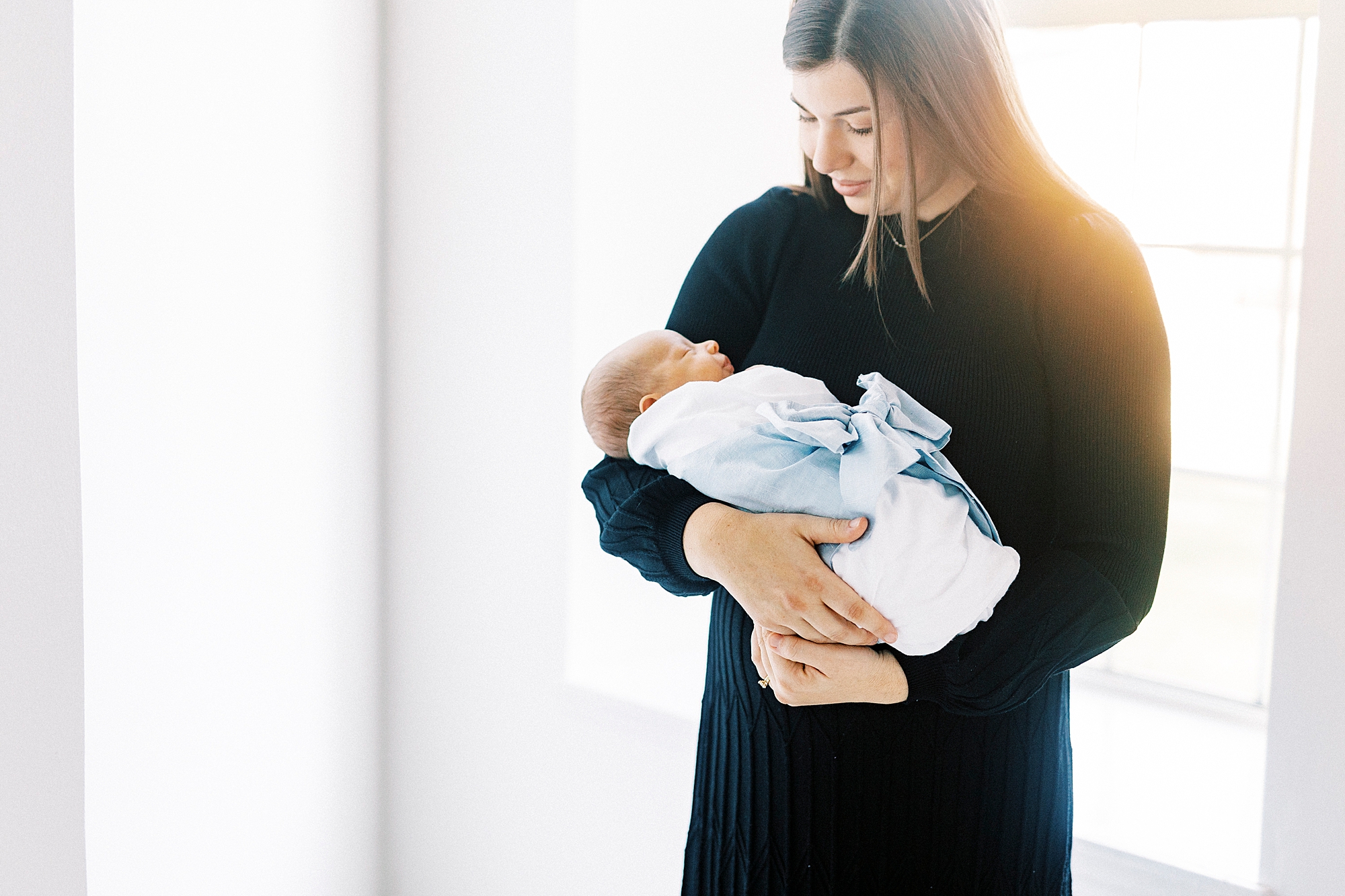 woman in black dress looks down at baby boy standing in window of Concord NC studio 
