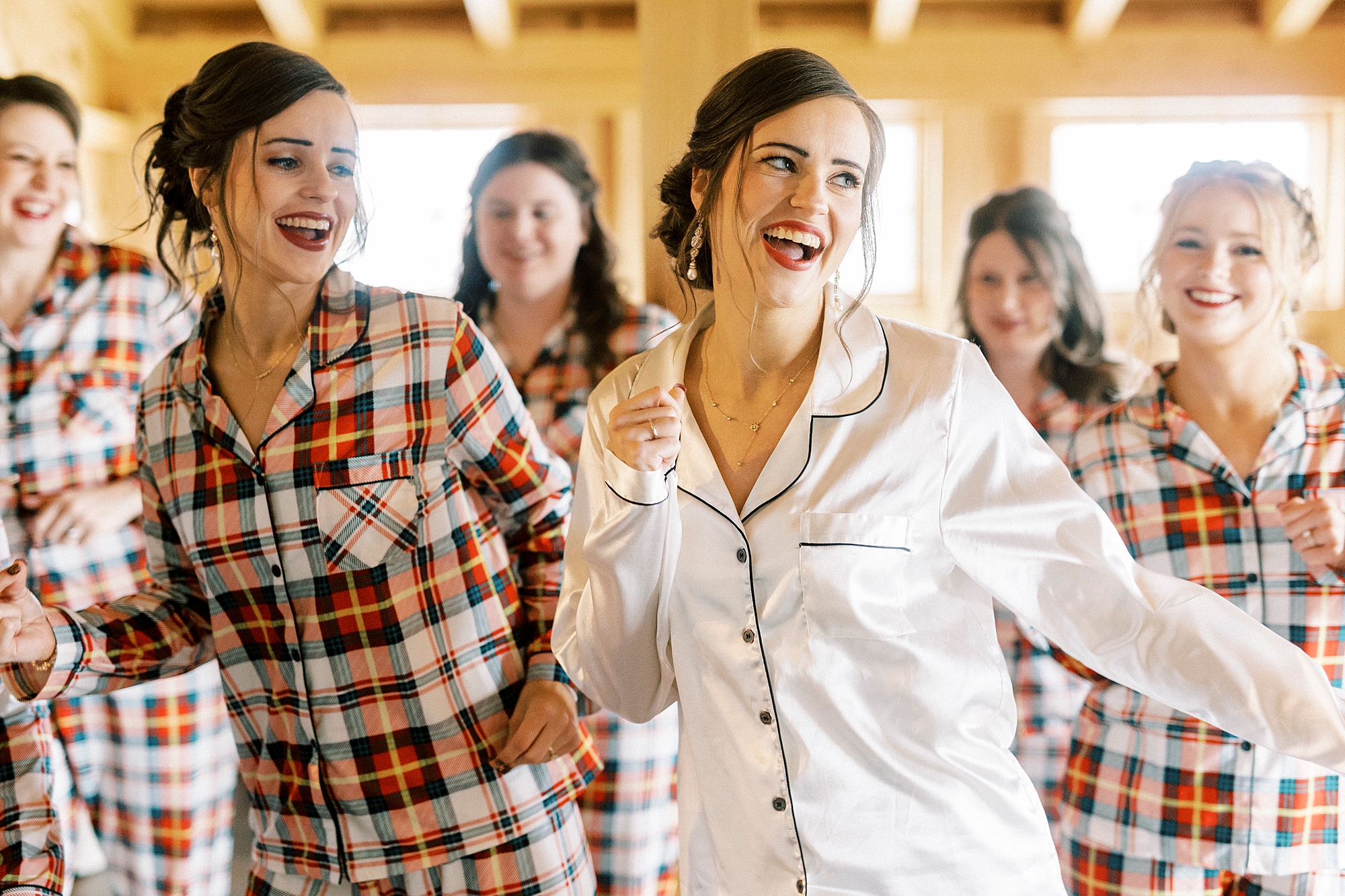 bride in white satin pajamas dances with bridesmaids in plaid pajamas before NC wedding