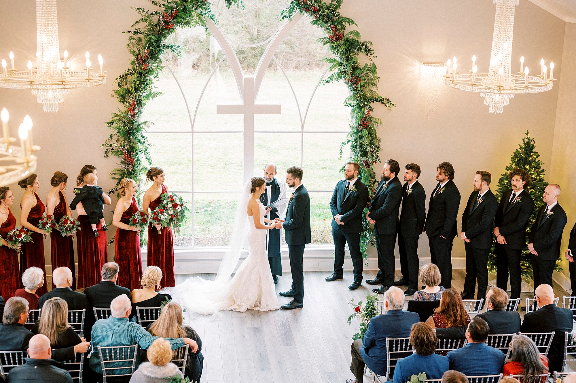 bride and groom hold hands in front of white cross and big windows for Chickadee Hill Farms wedding ceremony