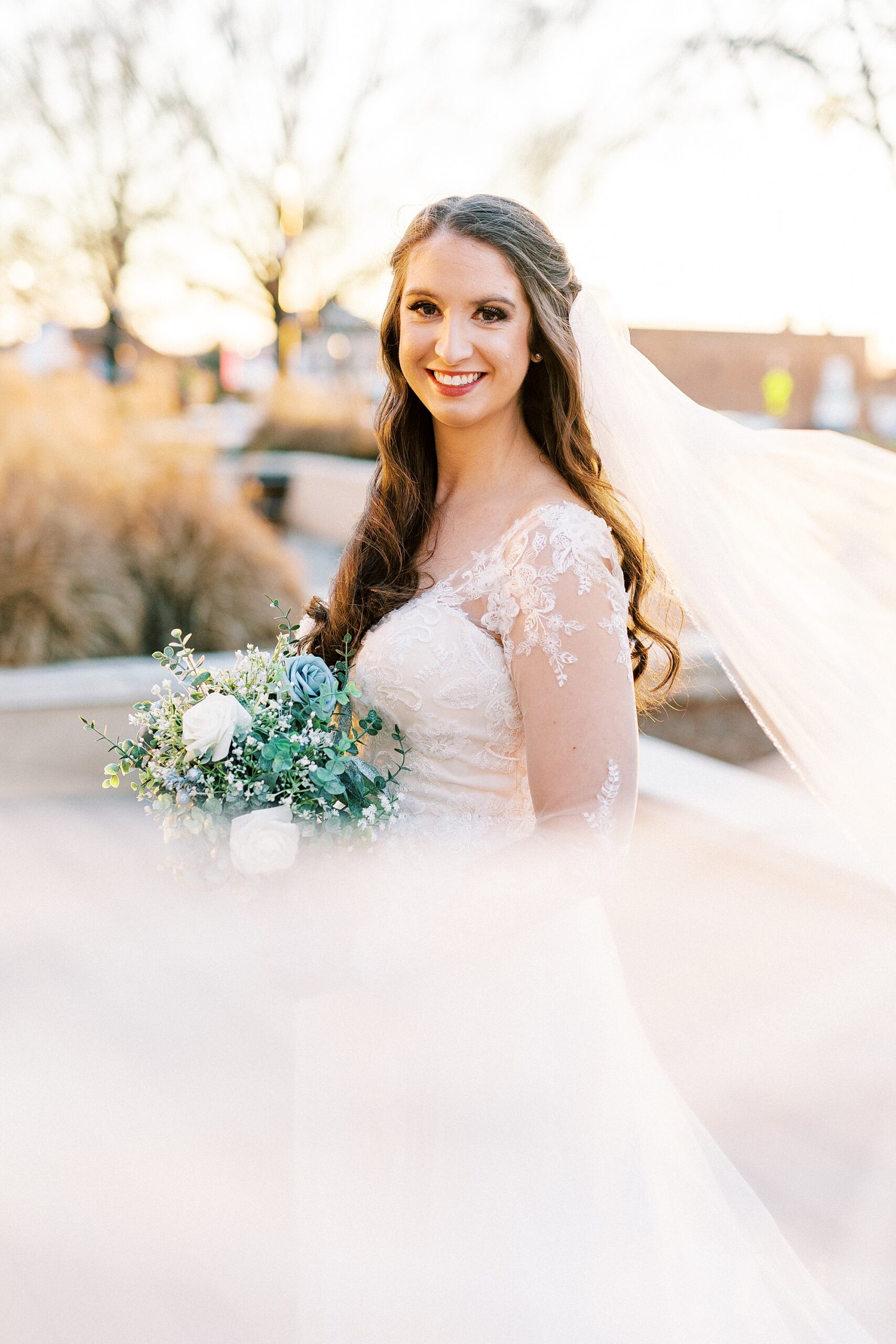 bride stands in wedding gown with lace sleeves and veil floating around her in Downtown Kannapolis
