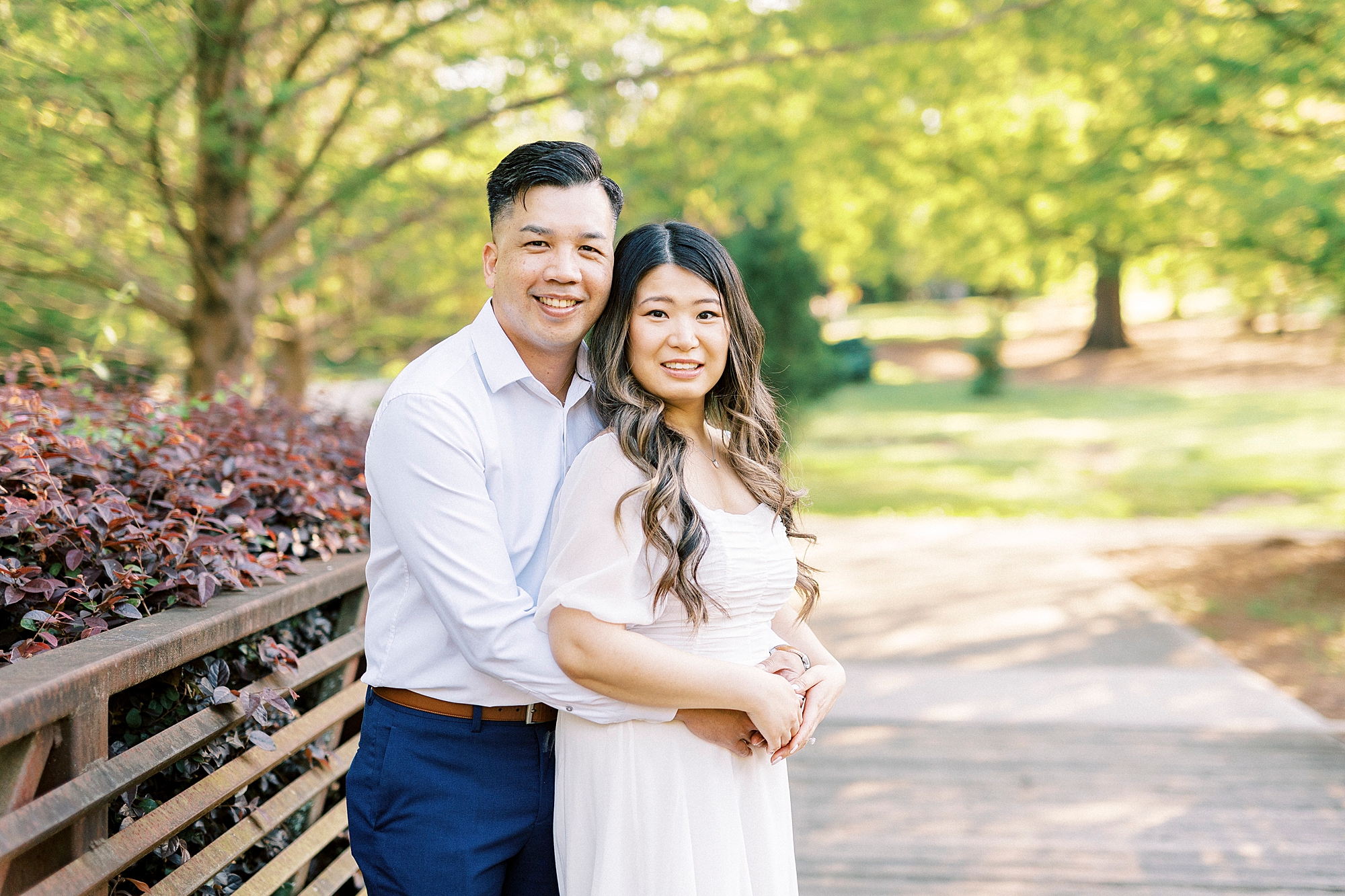 man hugs fiancee around waist from behind standing on a bridge 