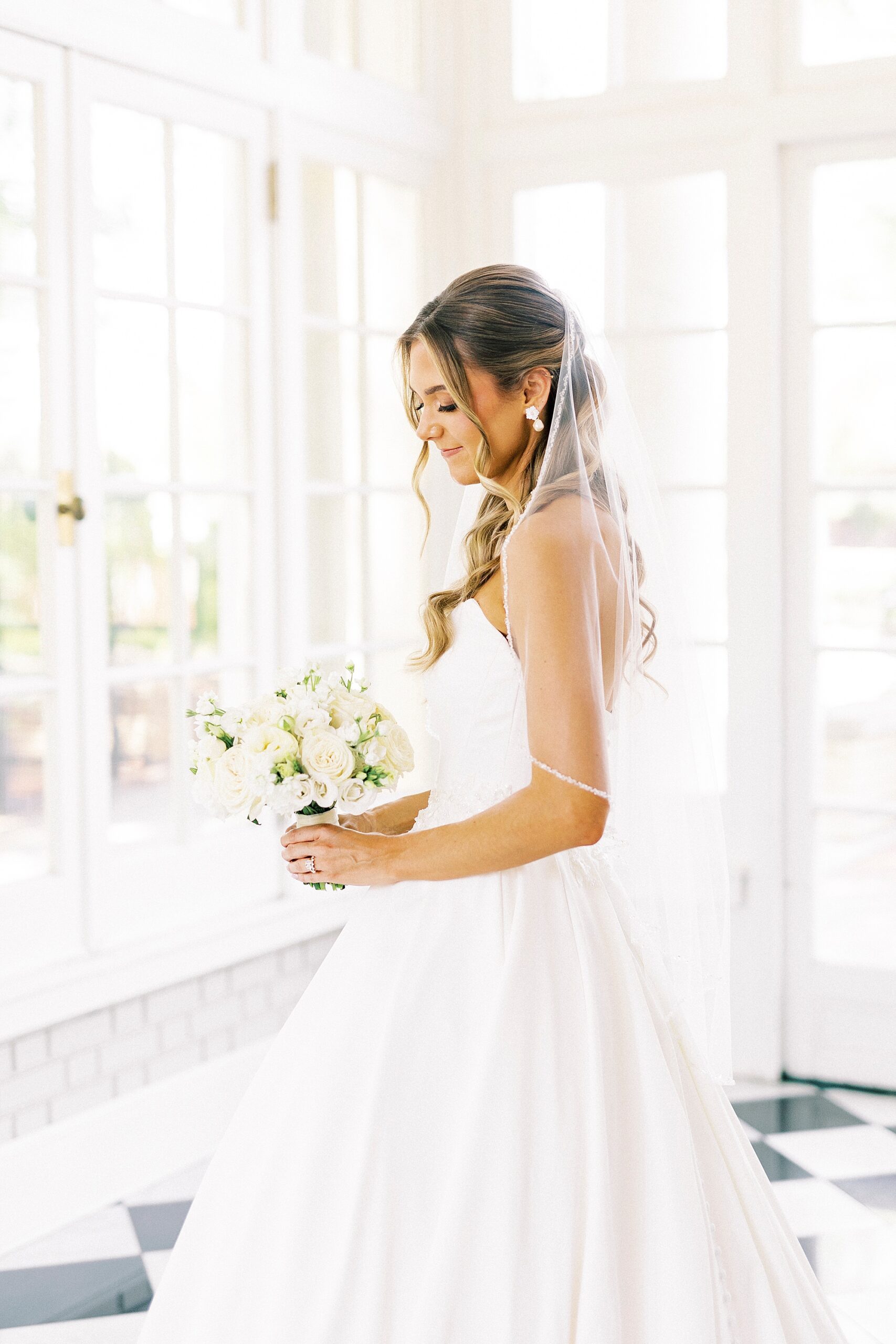 bride looks down at bouquet of ivory roses with veil around her shoulders 