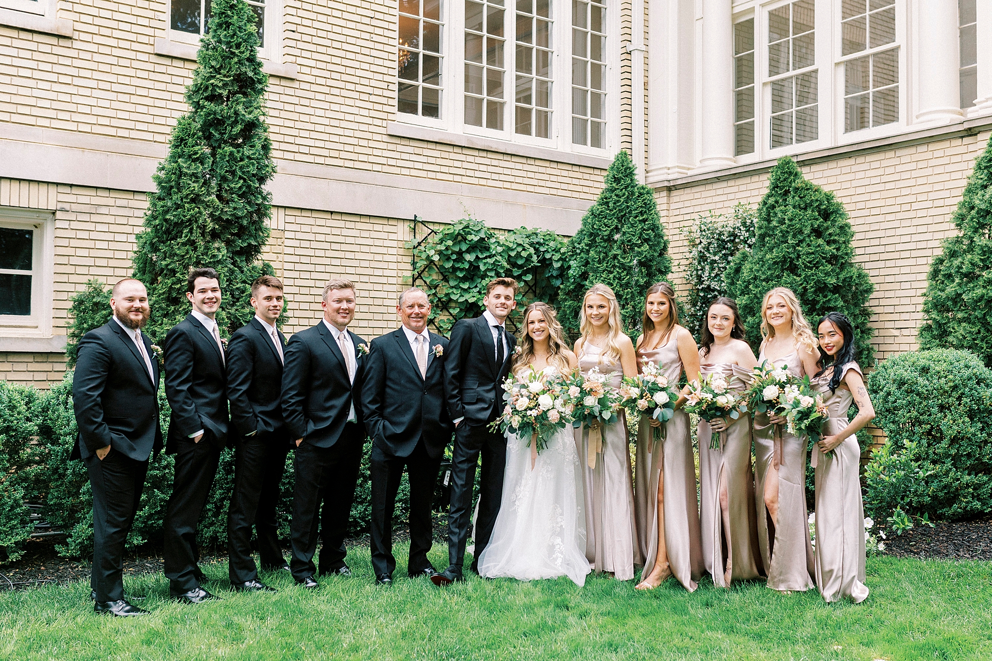wedding party in pink dresses and black suits stand with bride and groom on lawn at Separk Mansion