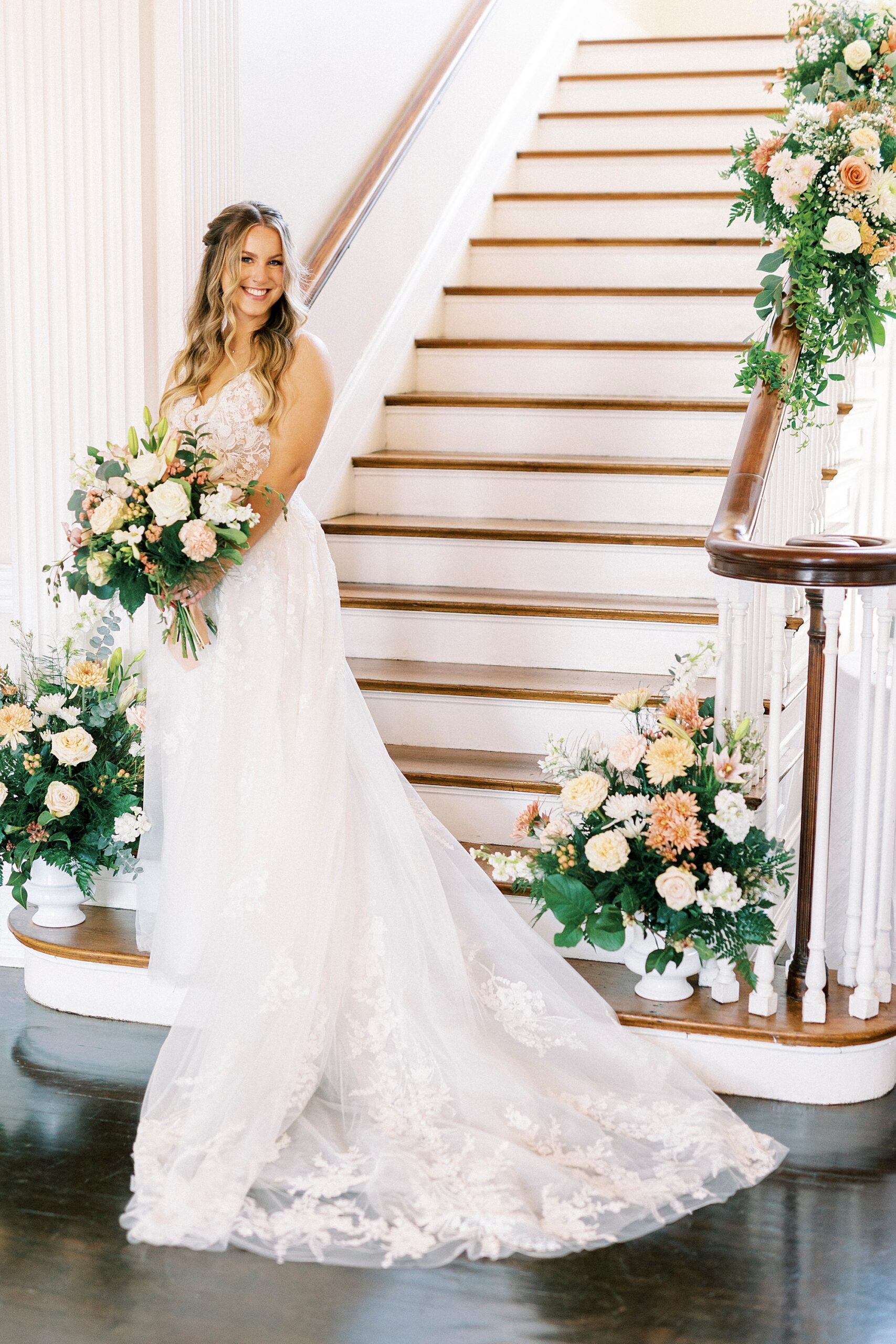 bride stands at bottom of steps holding bouquet of ivory and pink flowers inside Separk Mansion