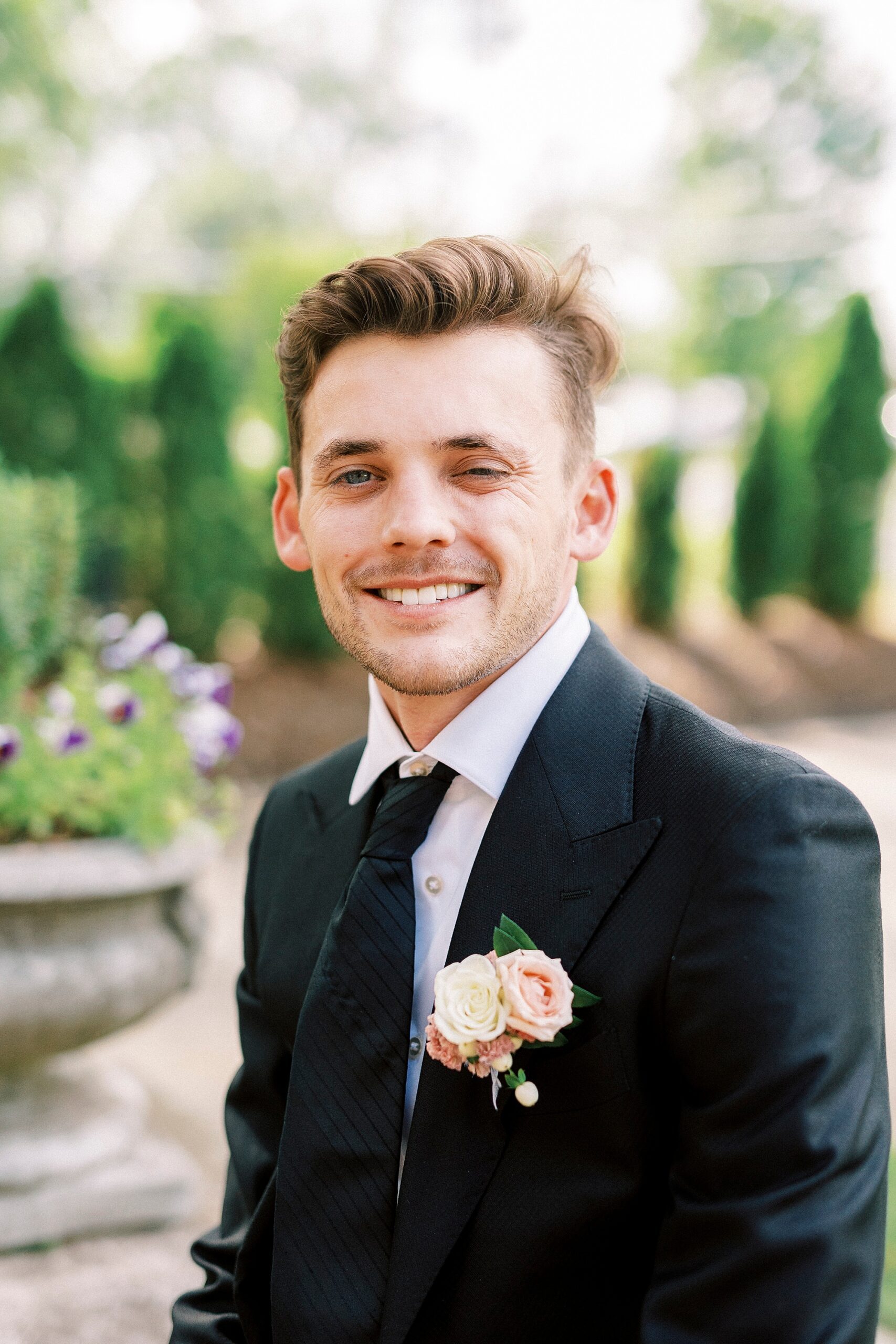 groom stands on brick patio with pink flower boutonnière 