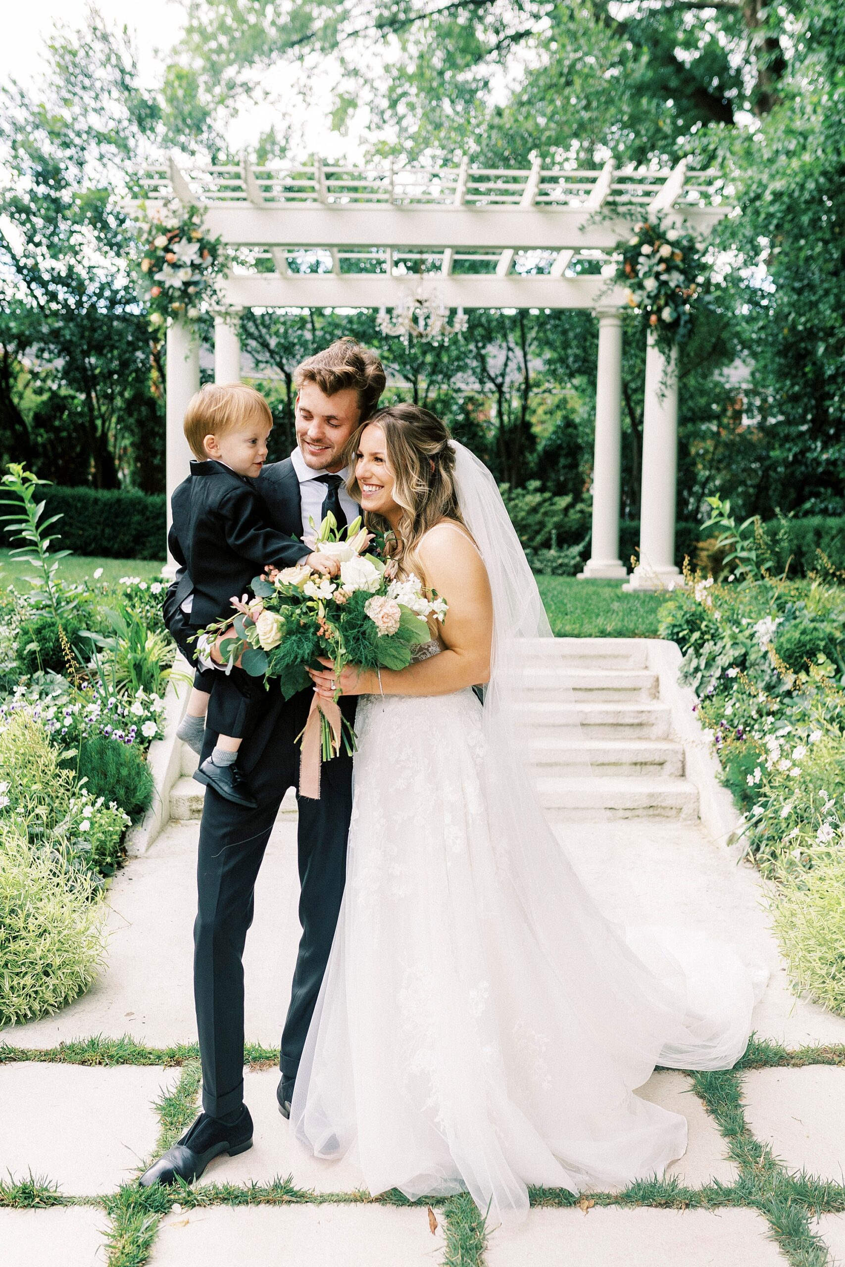 bride and groom smile in gardens of Separk Mansion with son on hip 