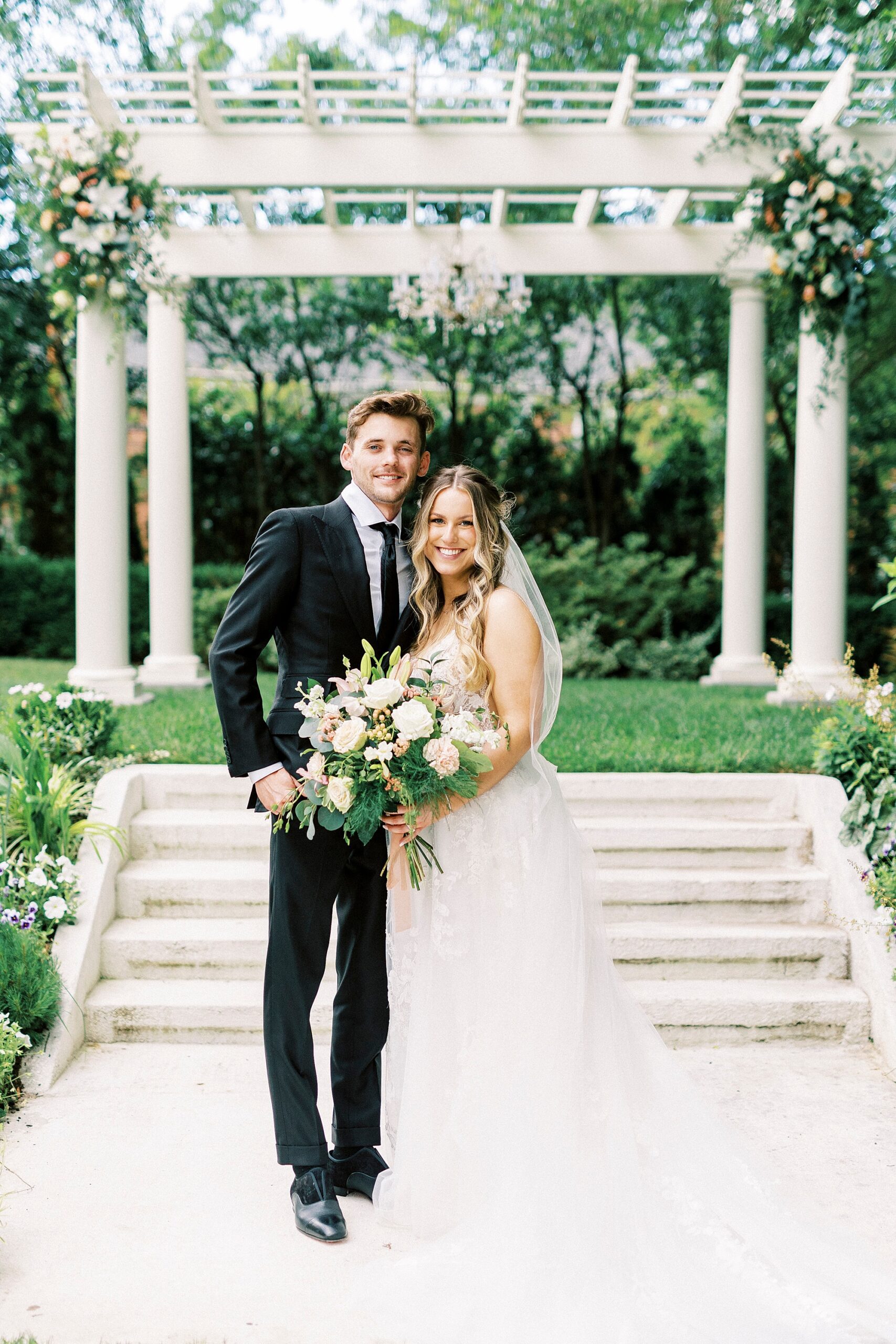 bride and groom stand in front of arbor in garden