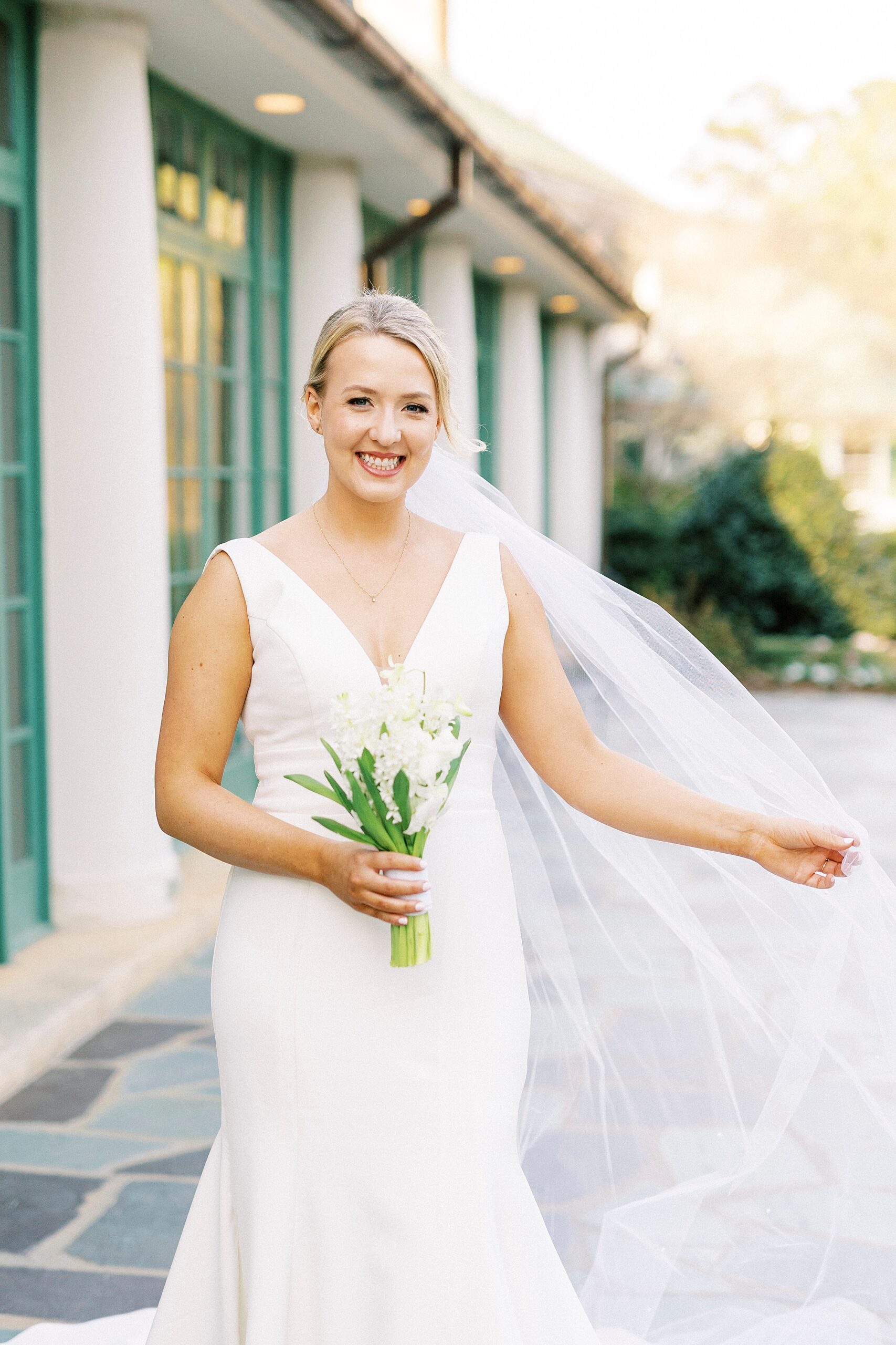 bride holds out veil around her standing by building at Reynolda Gardens