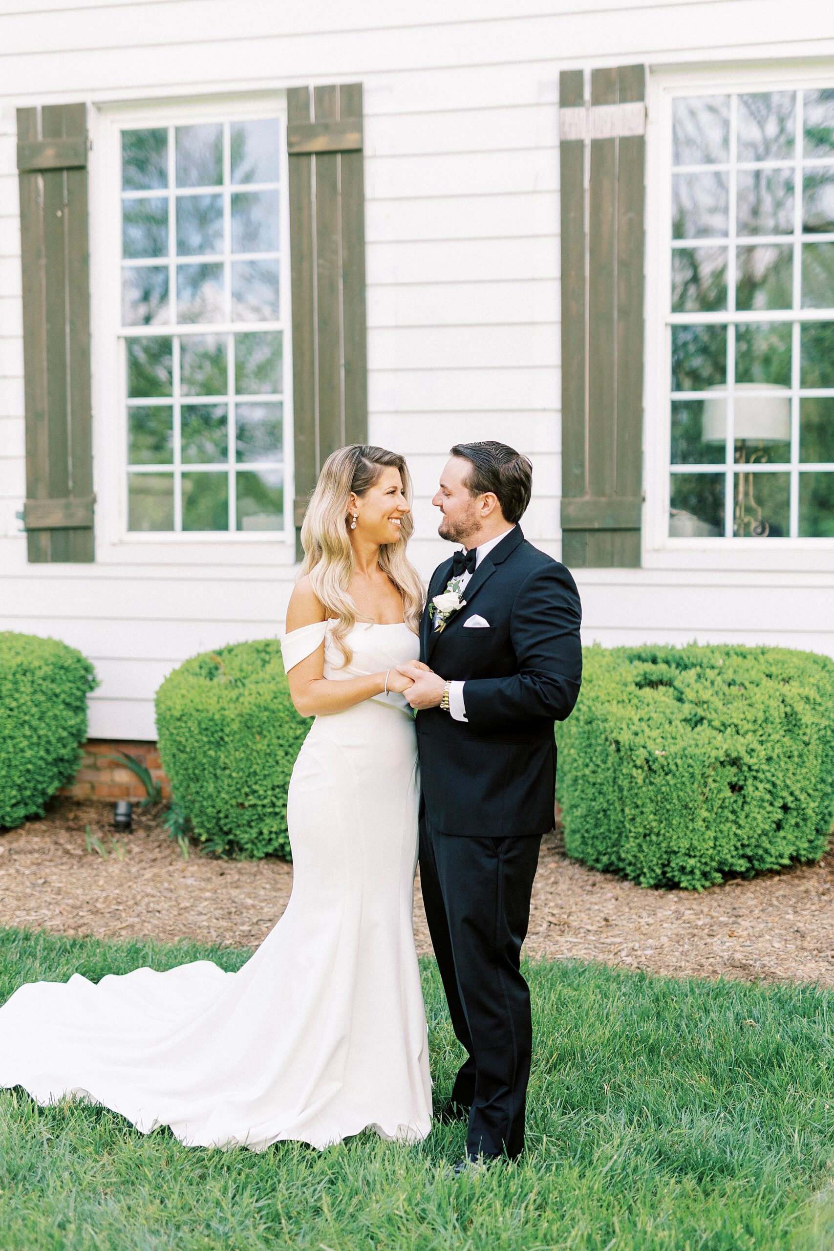bride and groom smile at each other in front of white house with green shutters