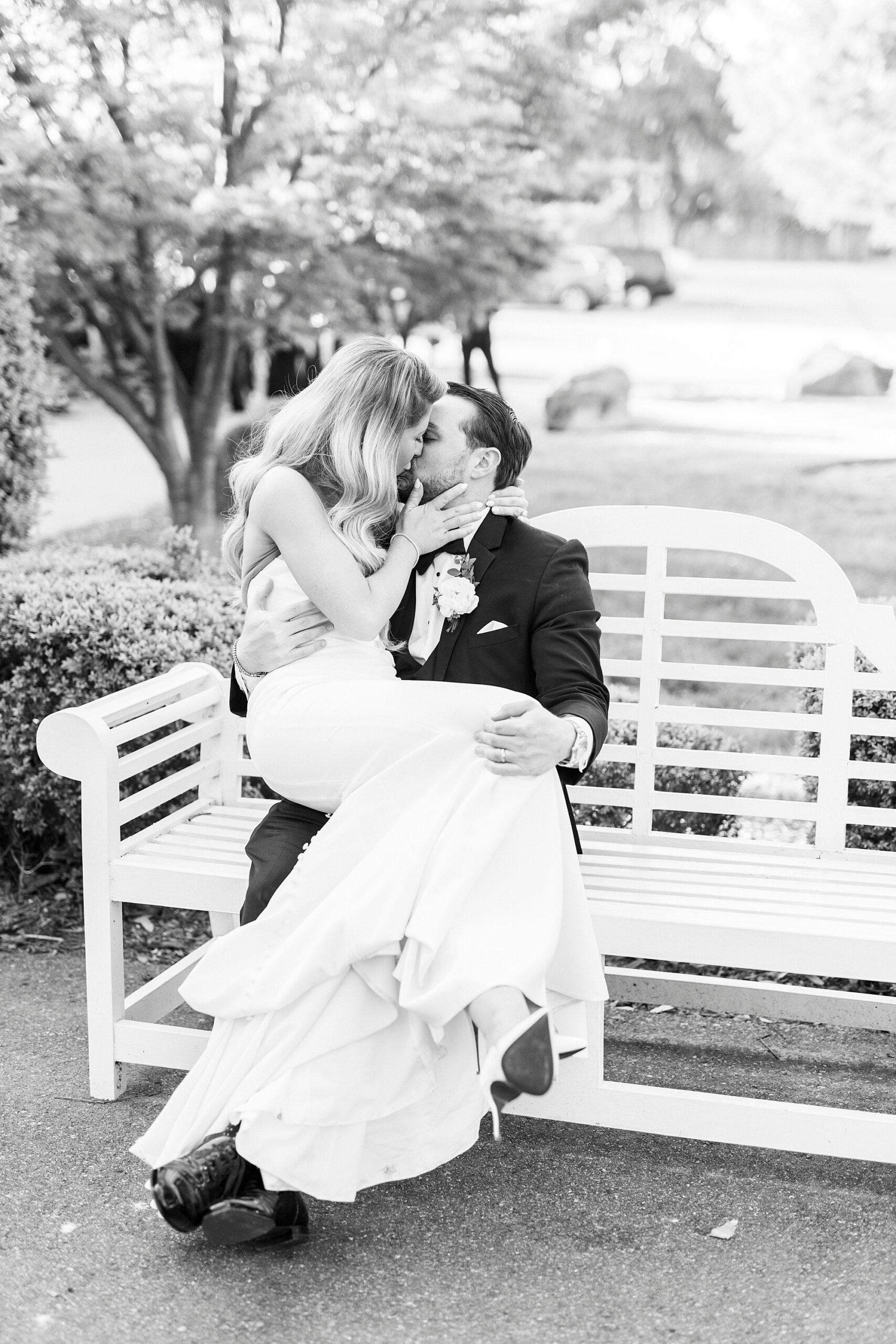 bride sits on groom's lap on white bench kissing
