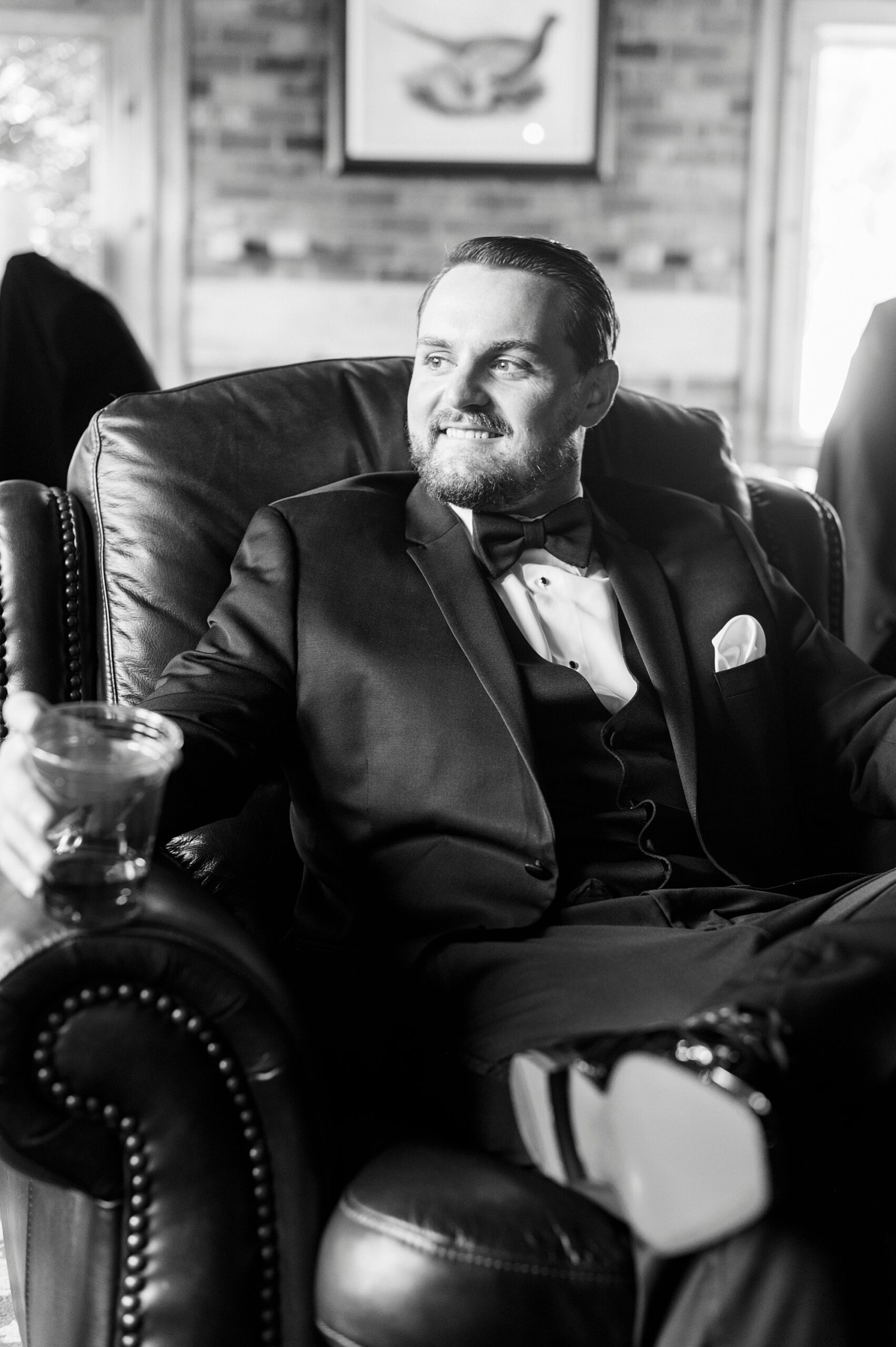 groom sits in leather chair in black tux for spring wedding