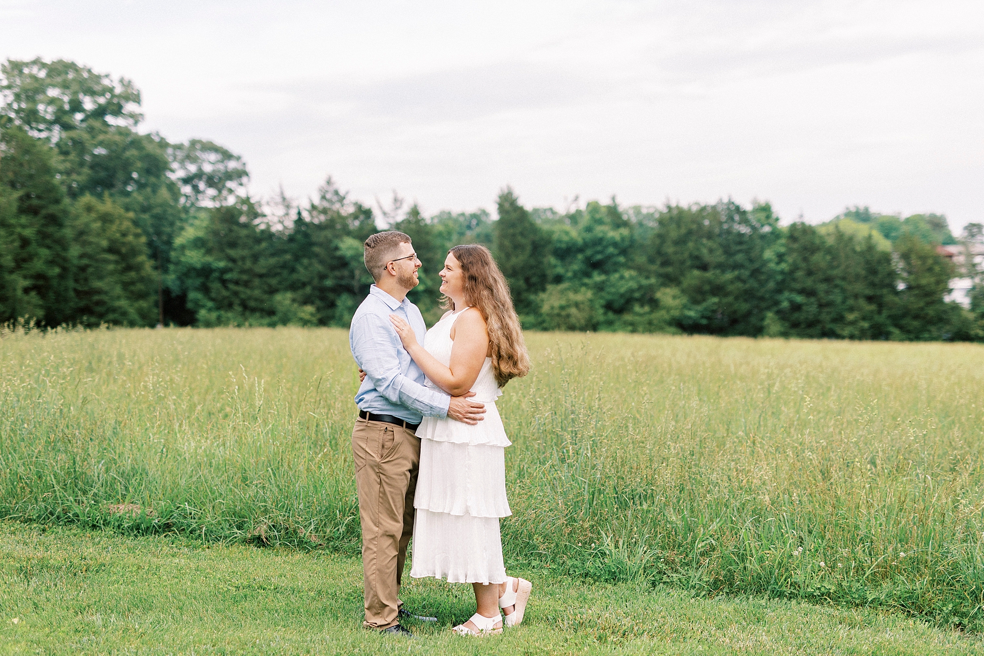 man hugs woman in white dress around waist near field of grass at Summerfield Farms