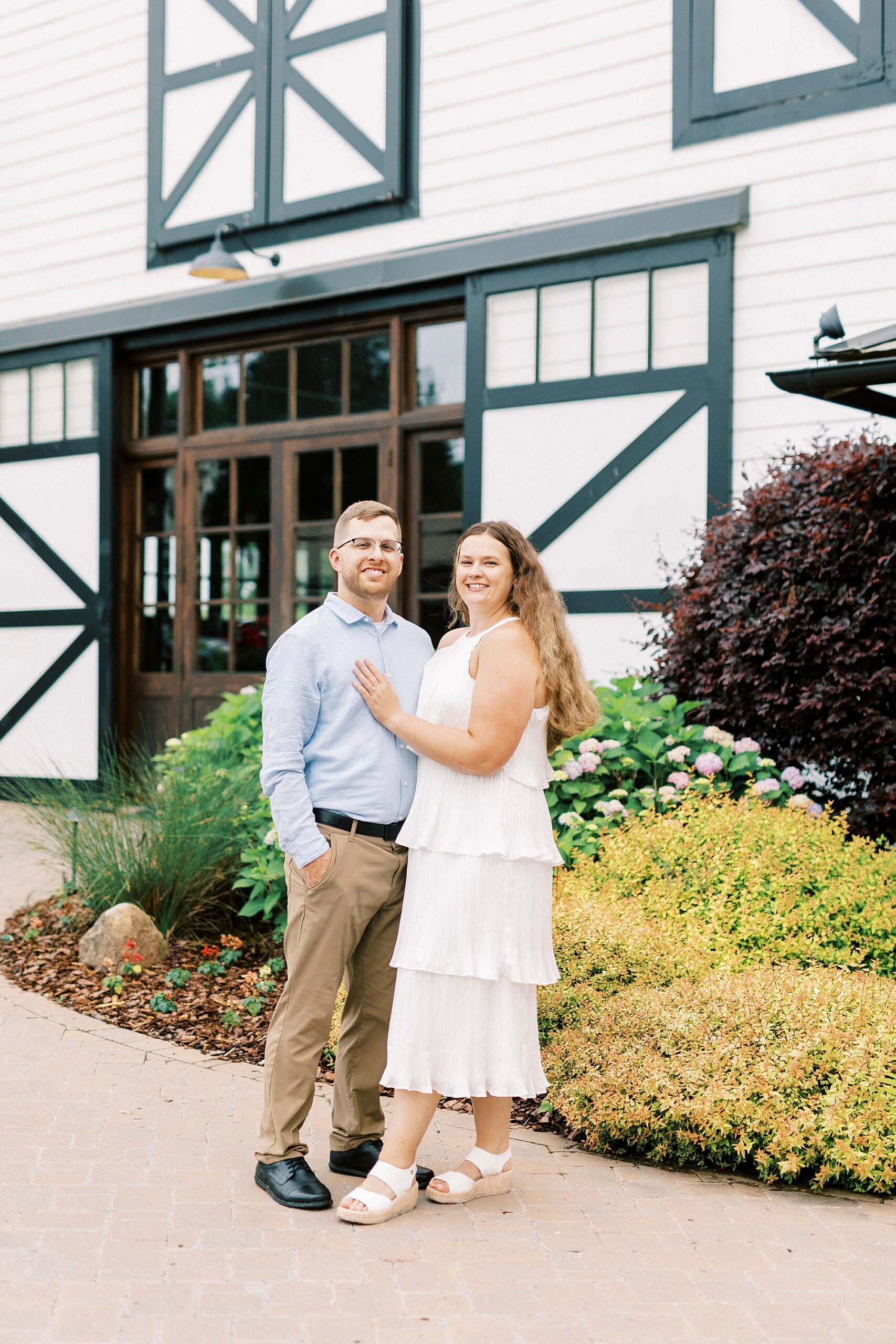 engaged couple stands by barn doors of building at Summerfield Farms
