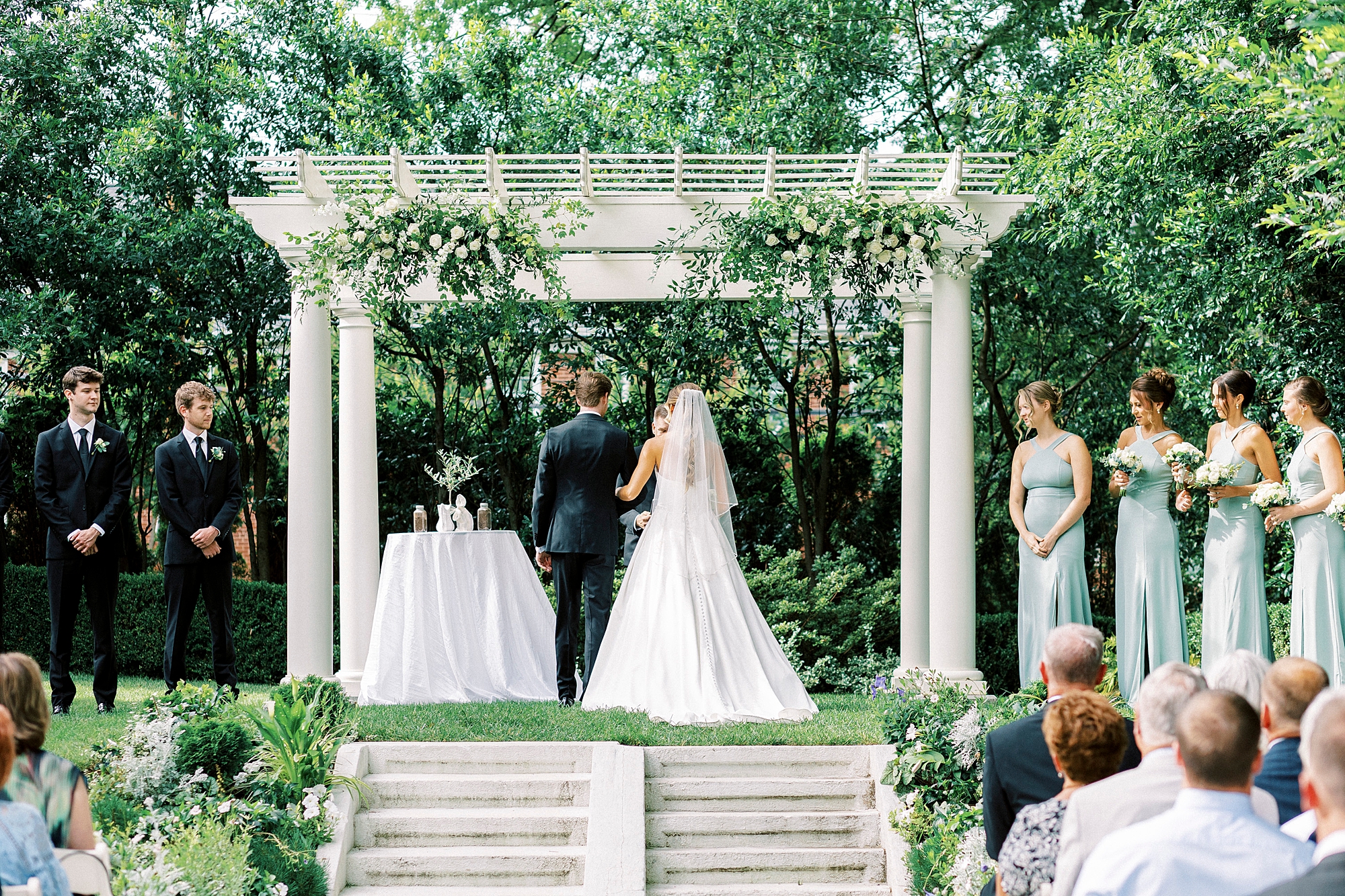 bride stands linking arms with groom during ceremony in gardens of Separk Mansion