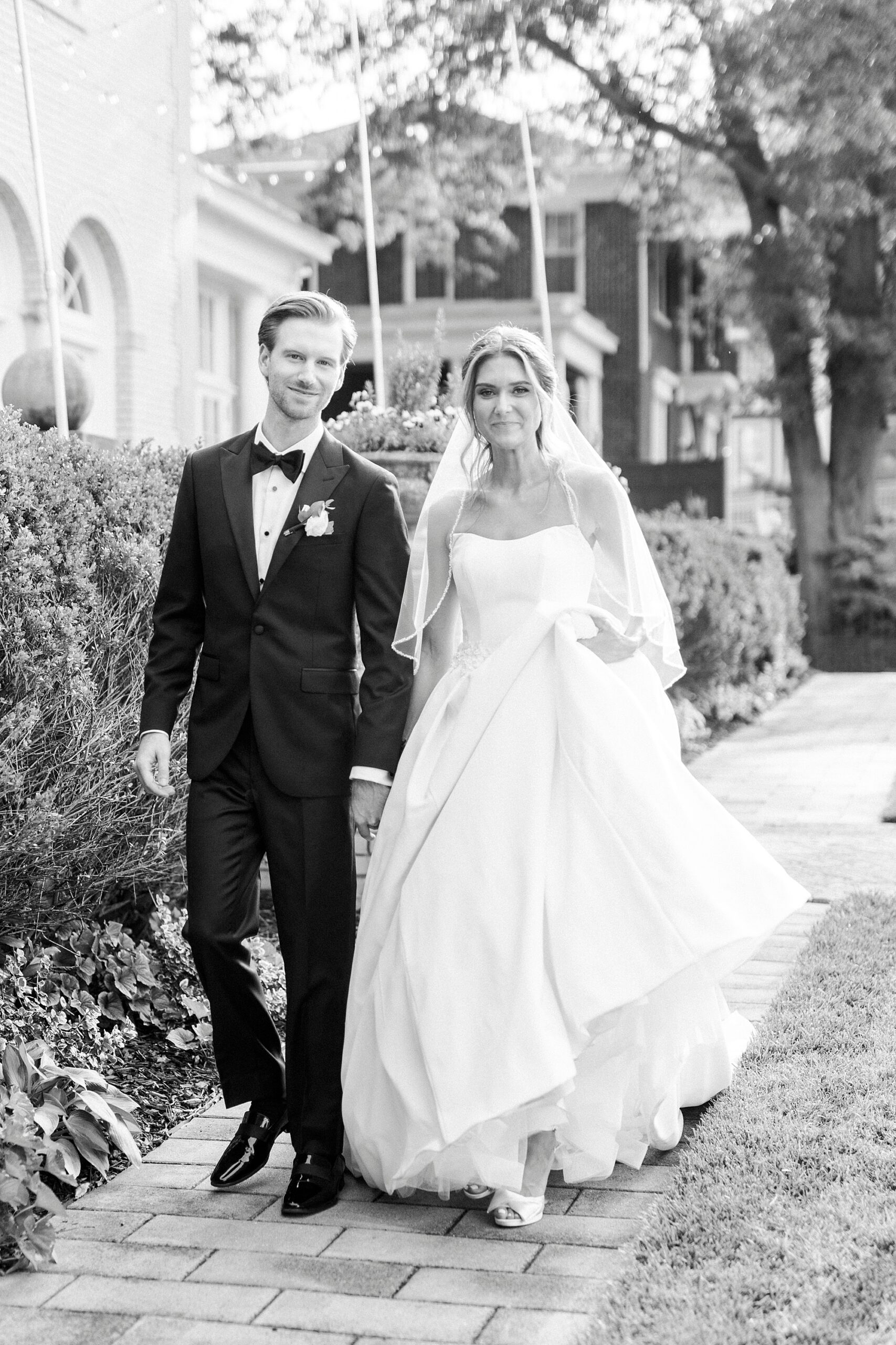 bride holds up skirt holding hands with groom walking through gardens 