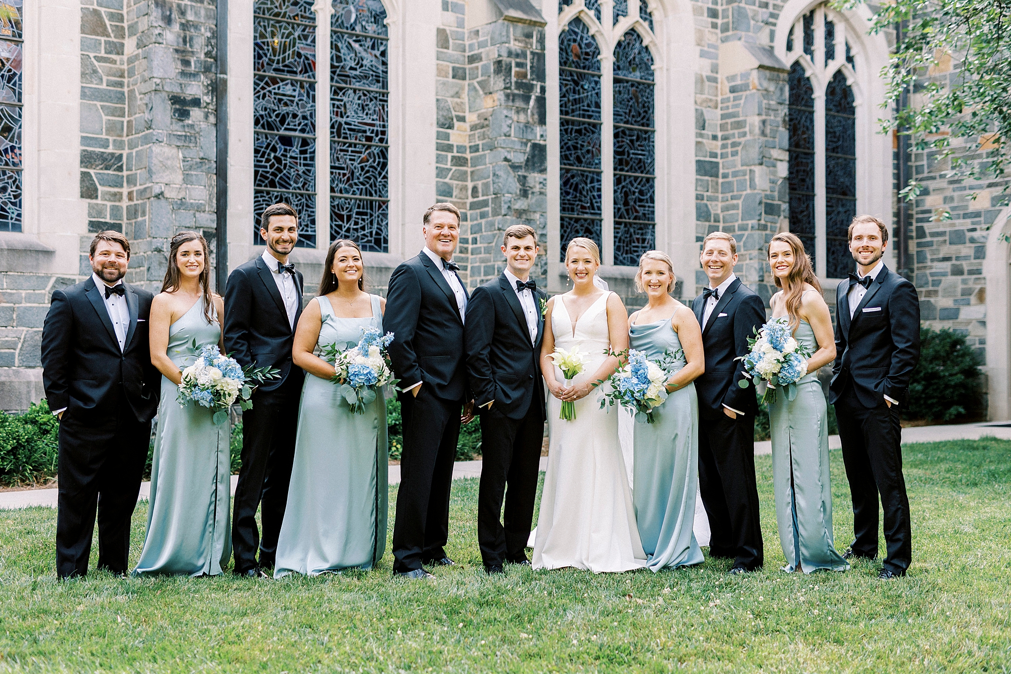 bride and groom stand with wedding party in black suits and blue gowns