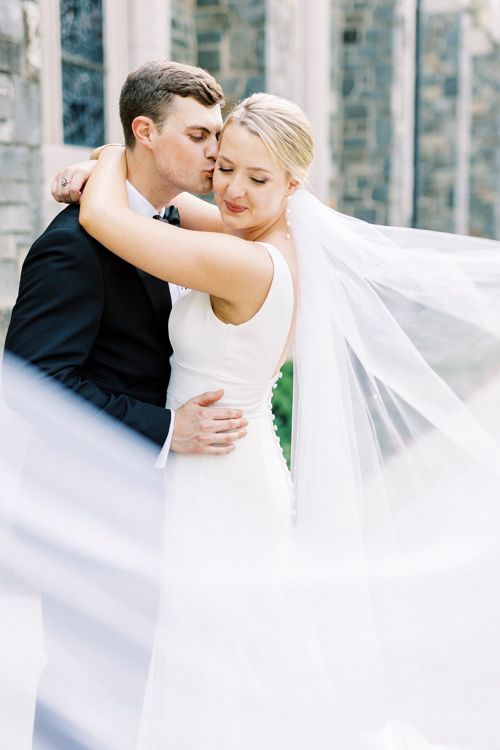 groom hugs bride kissing her cheek with veil around them