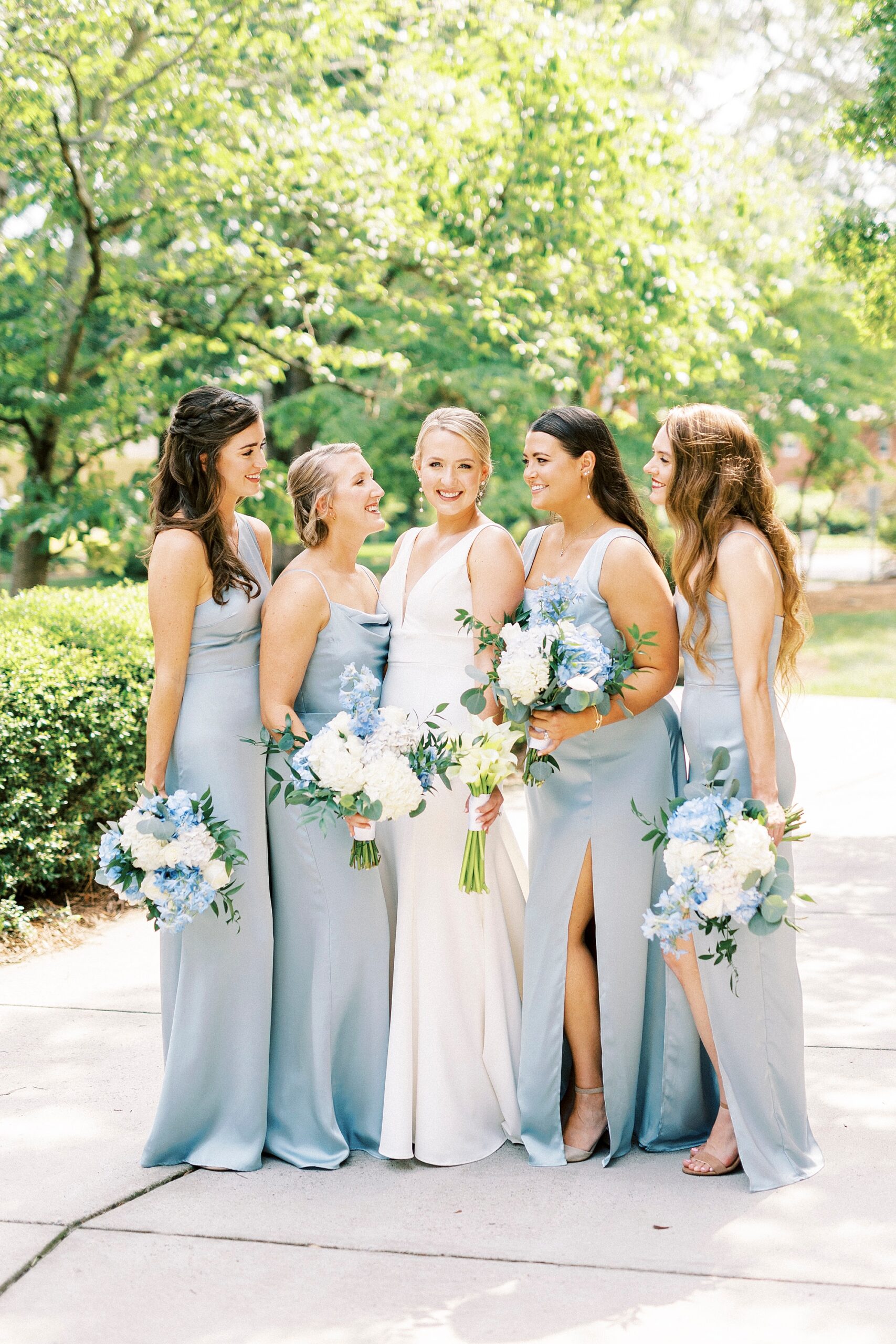 bride poses with bridesmaids in light blue gowns holding bouquet of blue and white flowers