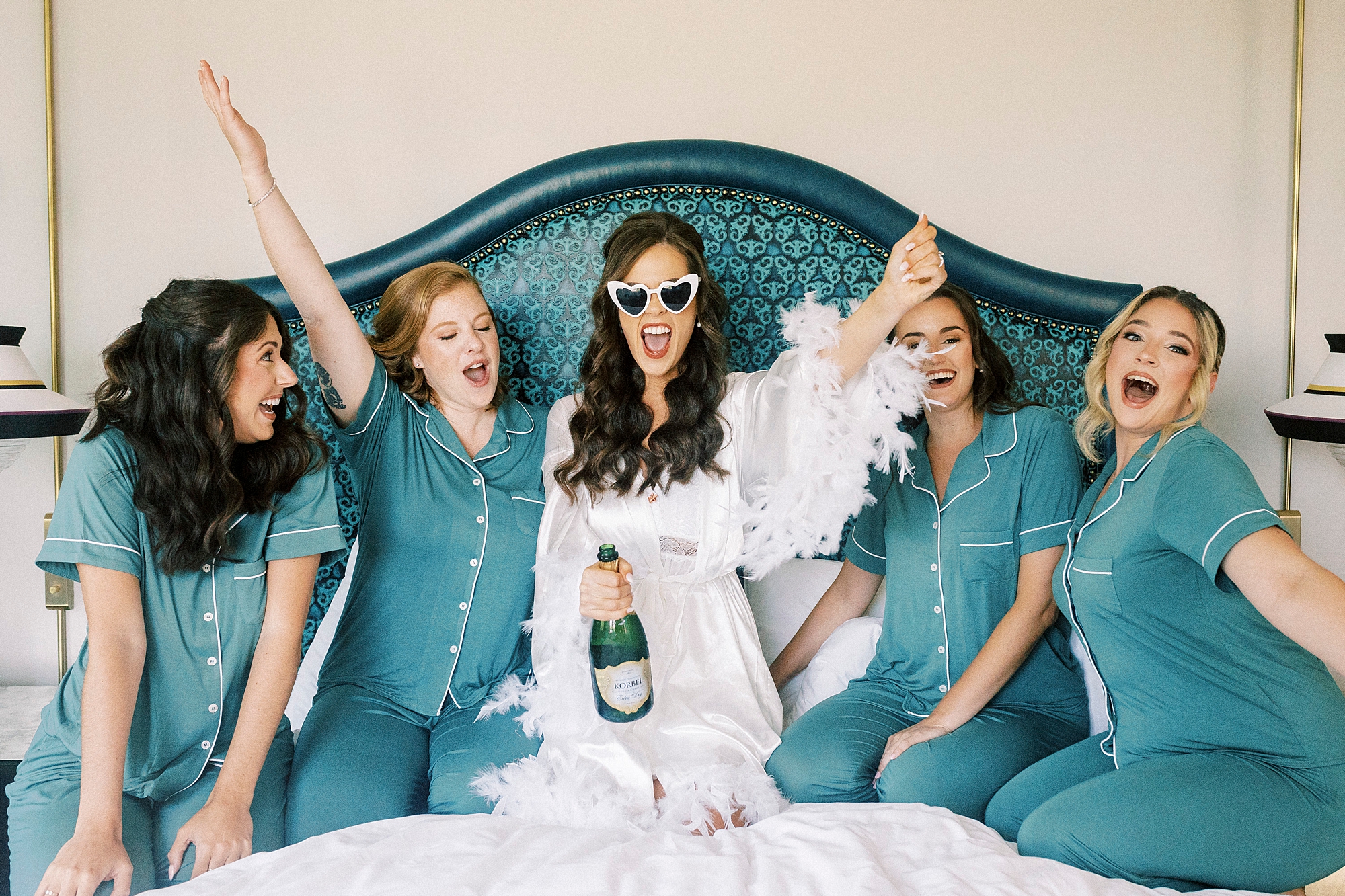 bride wears white sunglasses and robe with feathers sitting on bed cheering