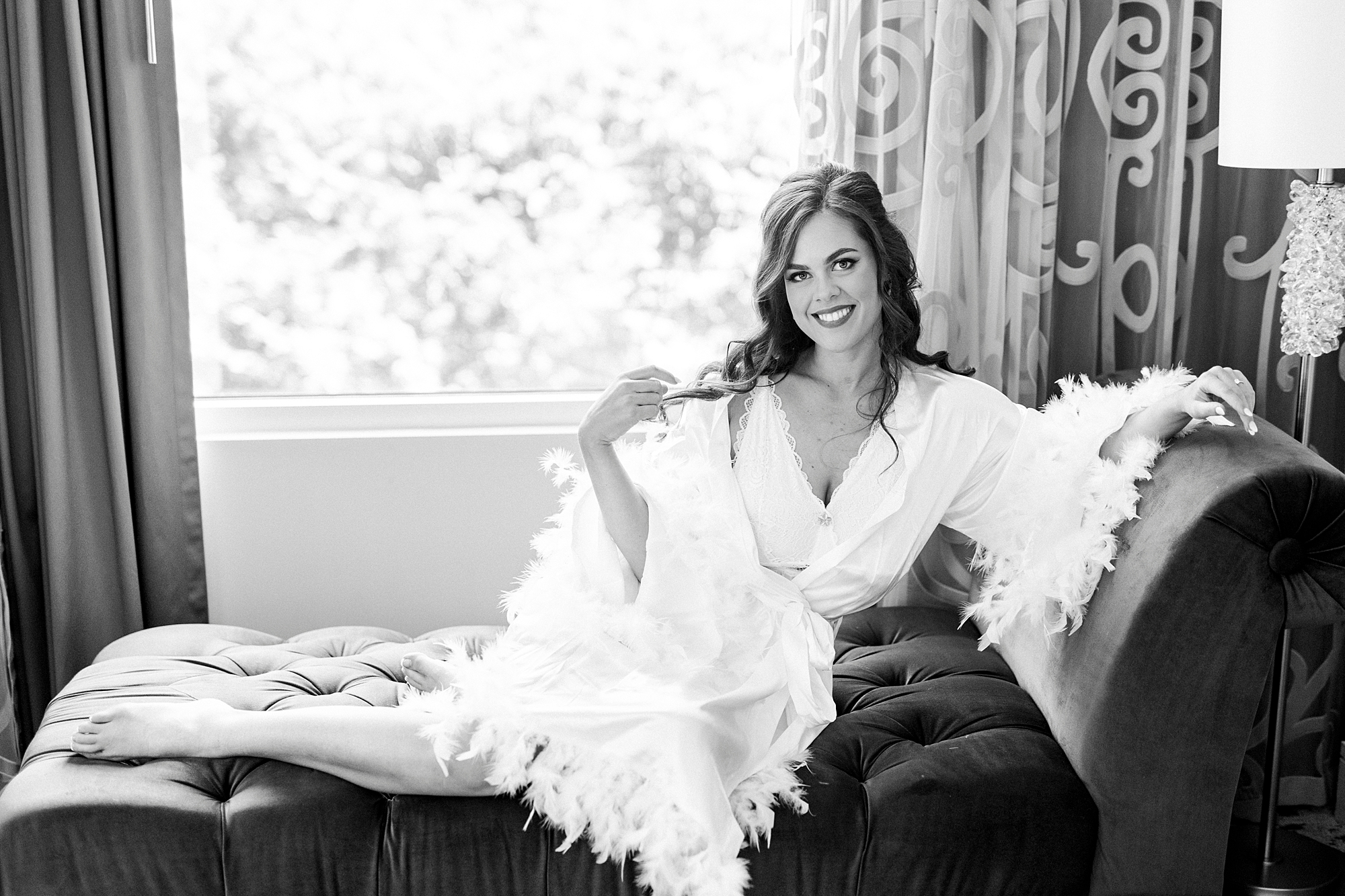 bride sits on lounge chair in feather robe during prep for Charlotte wedding