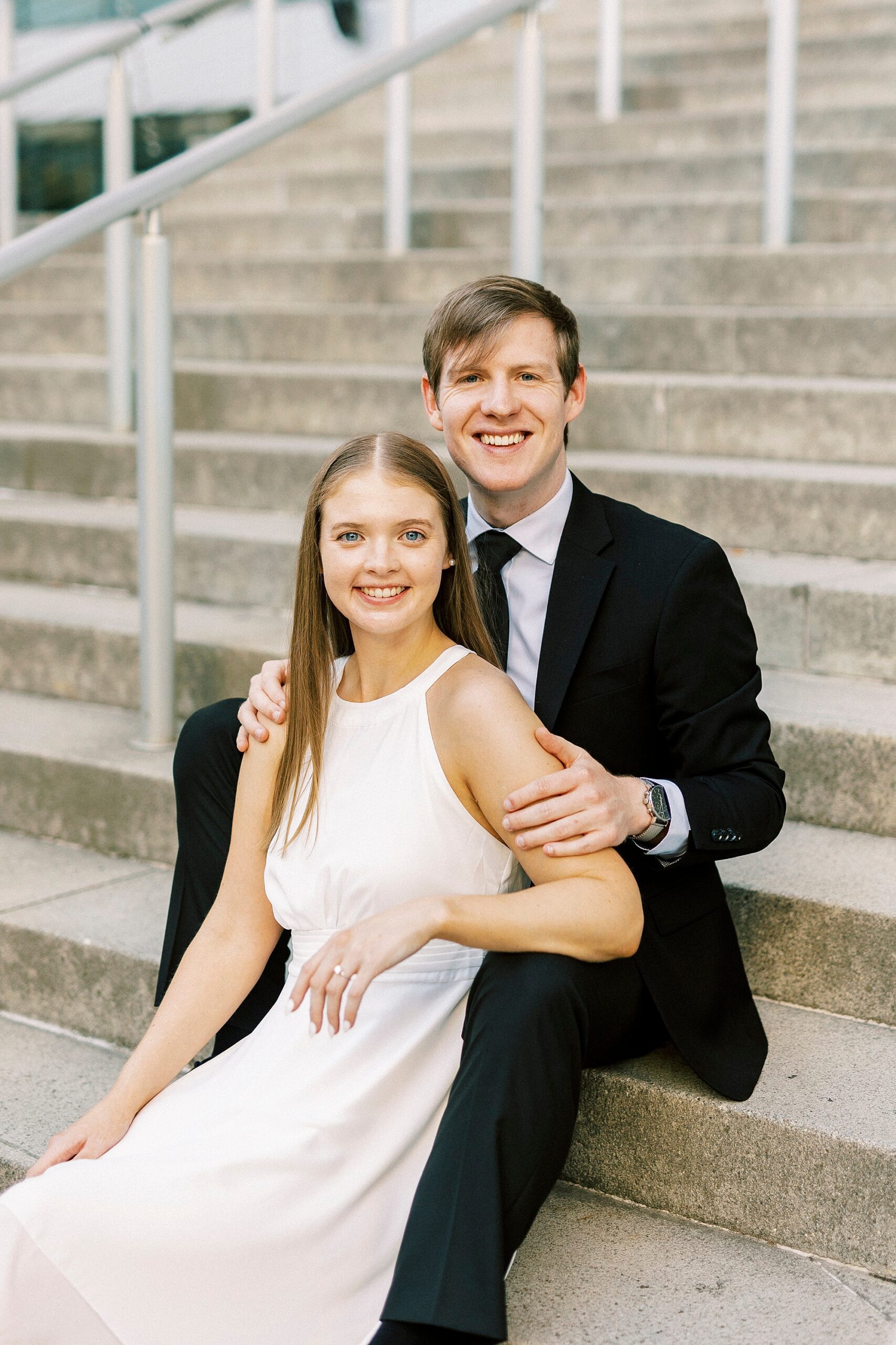 man smiles holding fiancee between his knees on steps 