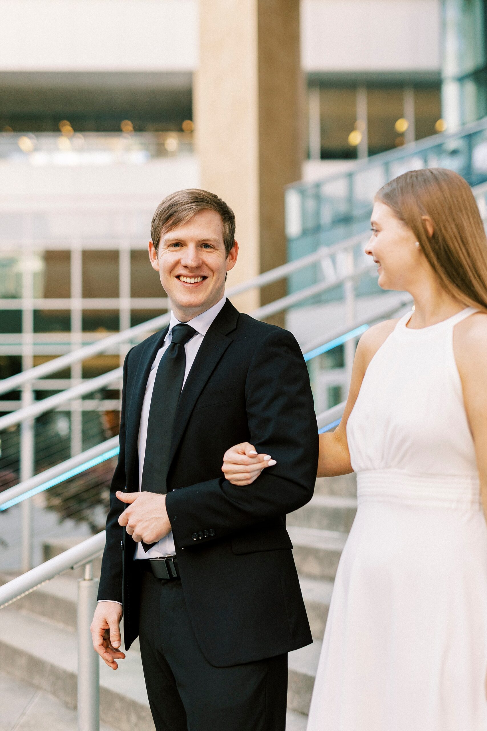 man in black suit escorts woman in white sundress down steps 