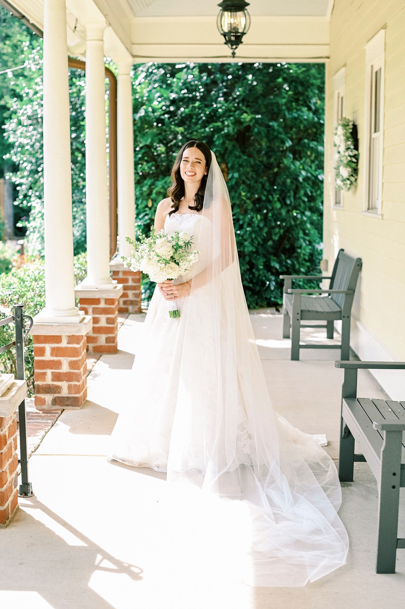 bride in strapless gown stands holding bouquet of ivory flowers with veil around her shoulders