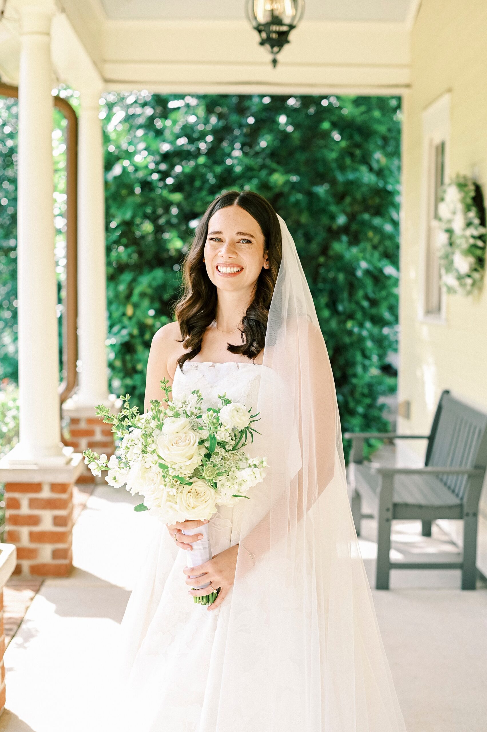 bride smiles holding bouquet of all white flowers on porch at Alexander Homestead