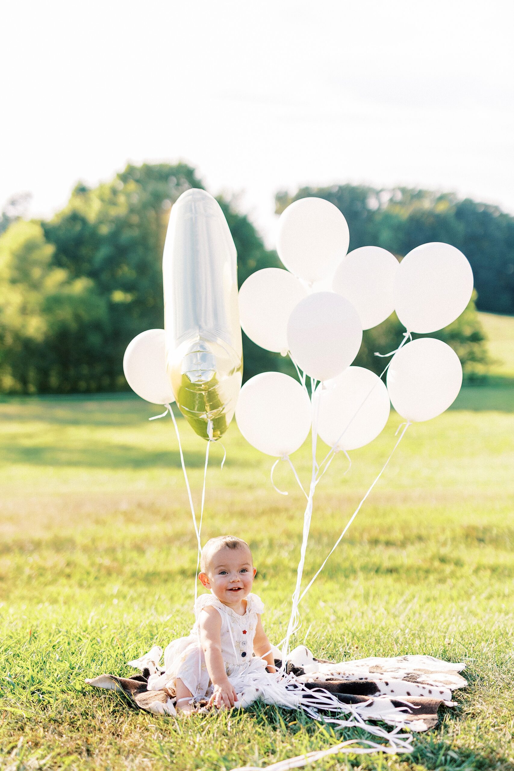toddler girl sits on blanket with white balloons during first birthday portraits in Albemarle NC