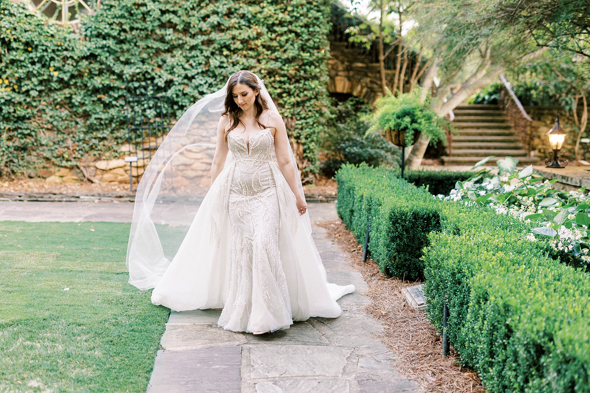 bride to be walks through gardens with veil trailing behind her